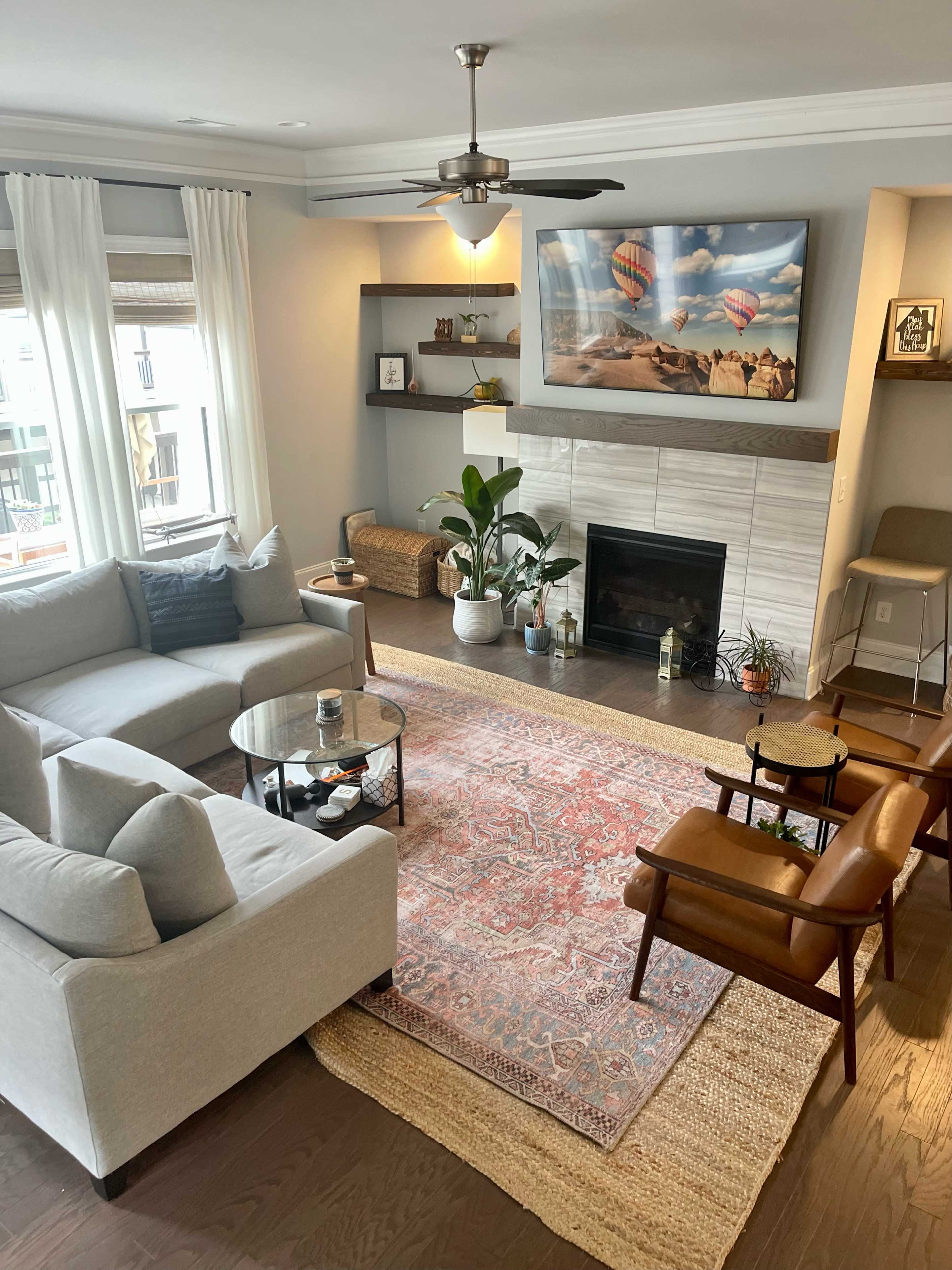A modern living room with a gray sectional sofa, a glass coffee table, and two brown lounge chairs, featuring a rug, plants, and a wall-mounted television above a fireplace.