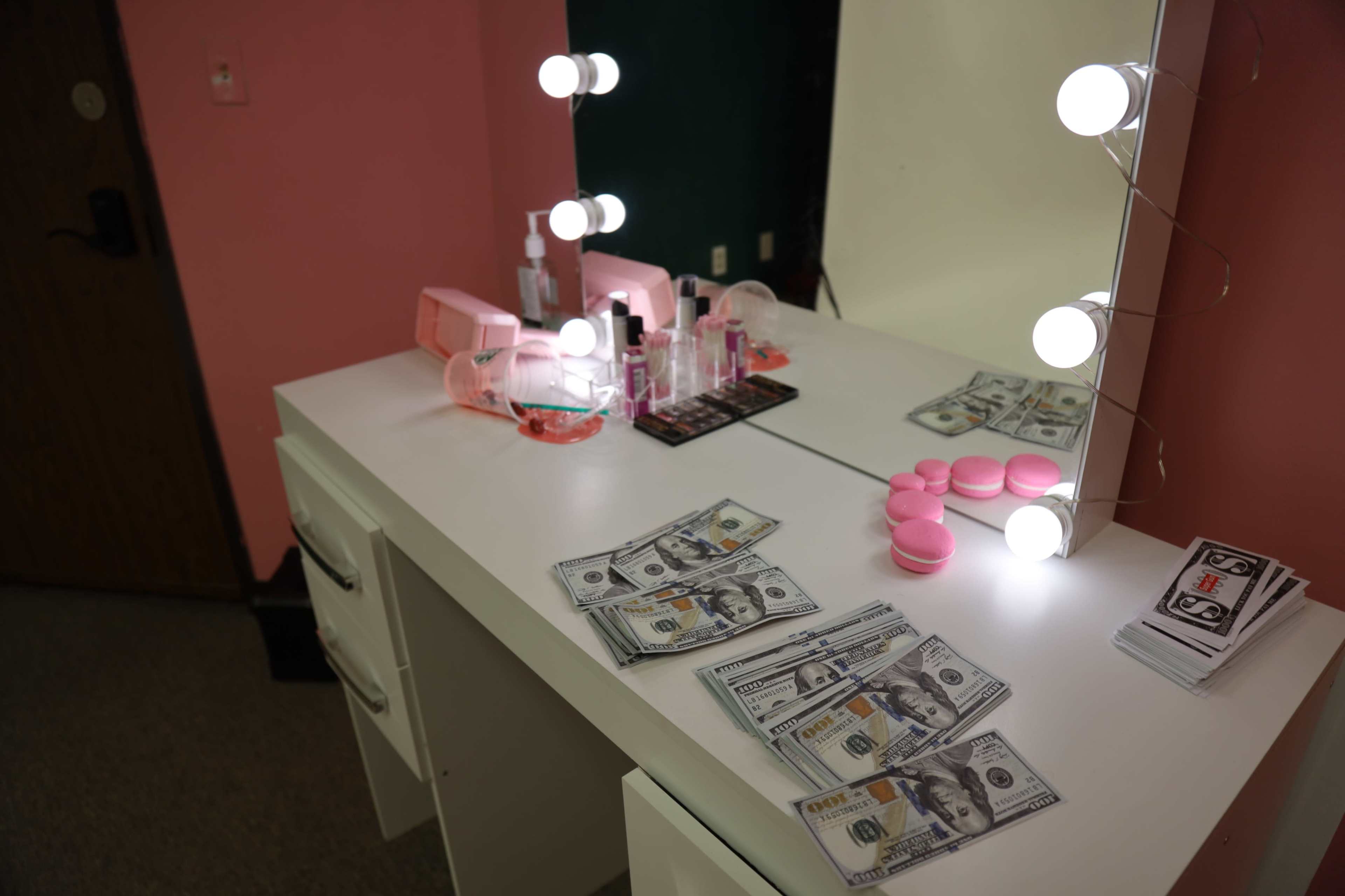 A vanity table is adorned with cosmetics, several stacks of cash, and a few pink beauty items, illuminated by round light bulbs.