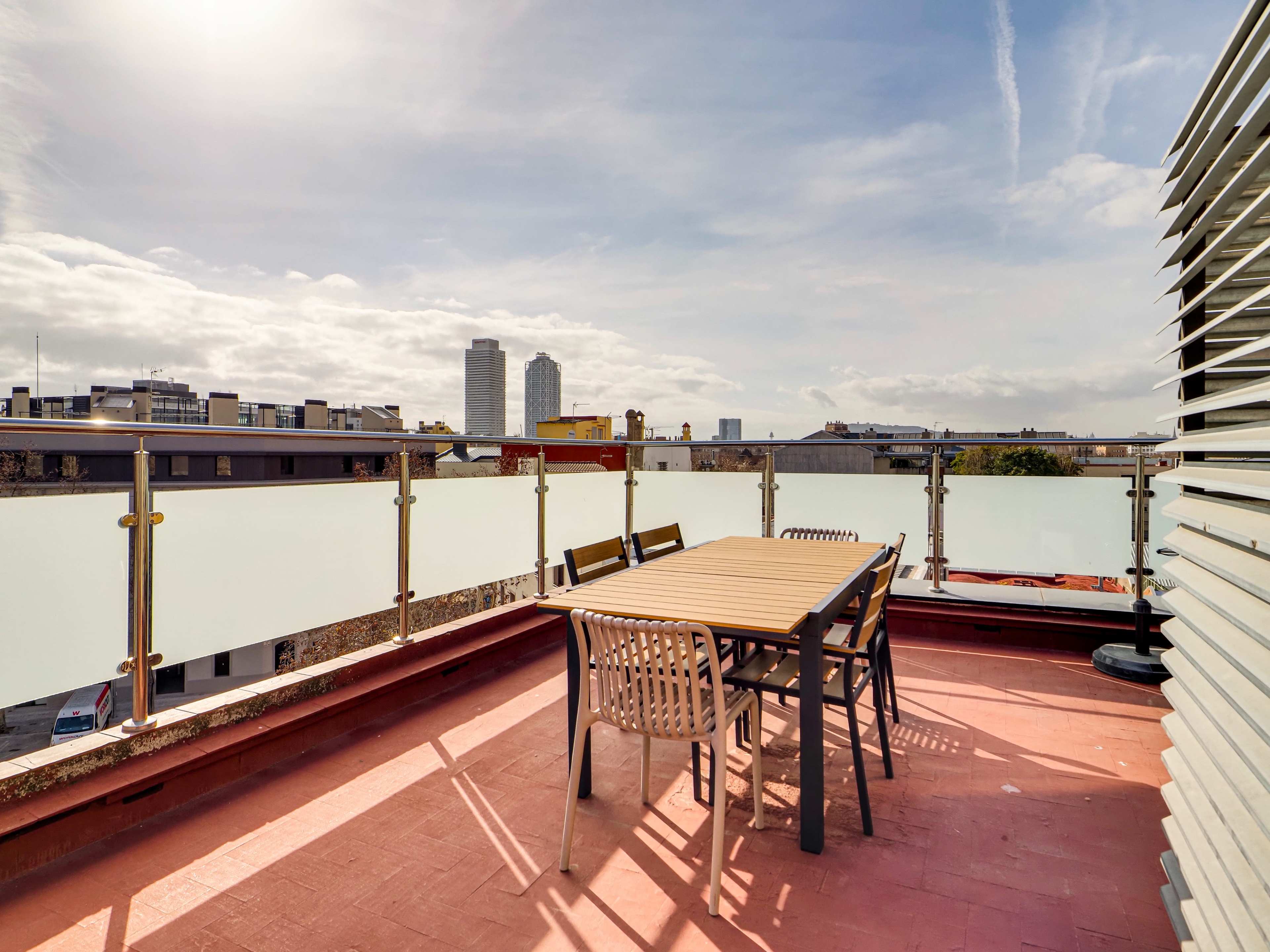 A rooftop terrace features a wooden table surrounded by chairs, with a view of tall buildings and a cloudy sky in the background.