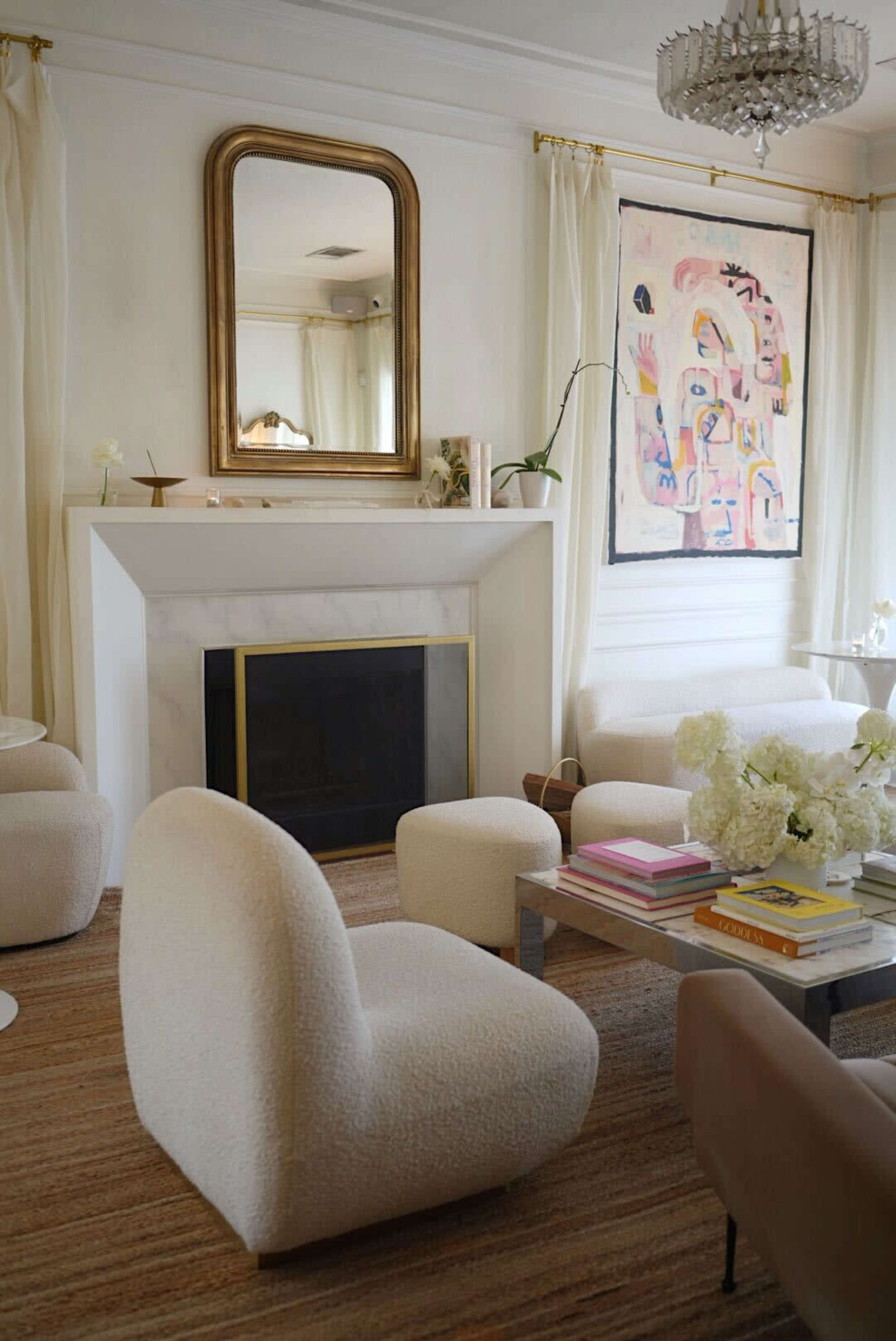 The image shows a modern living room featuring a fireplace, two cream-colored upholstered chairs, a glass coffee table with books and flowers, and a colorful abstract painting on the wall above the mantel.