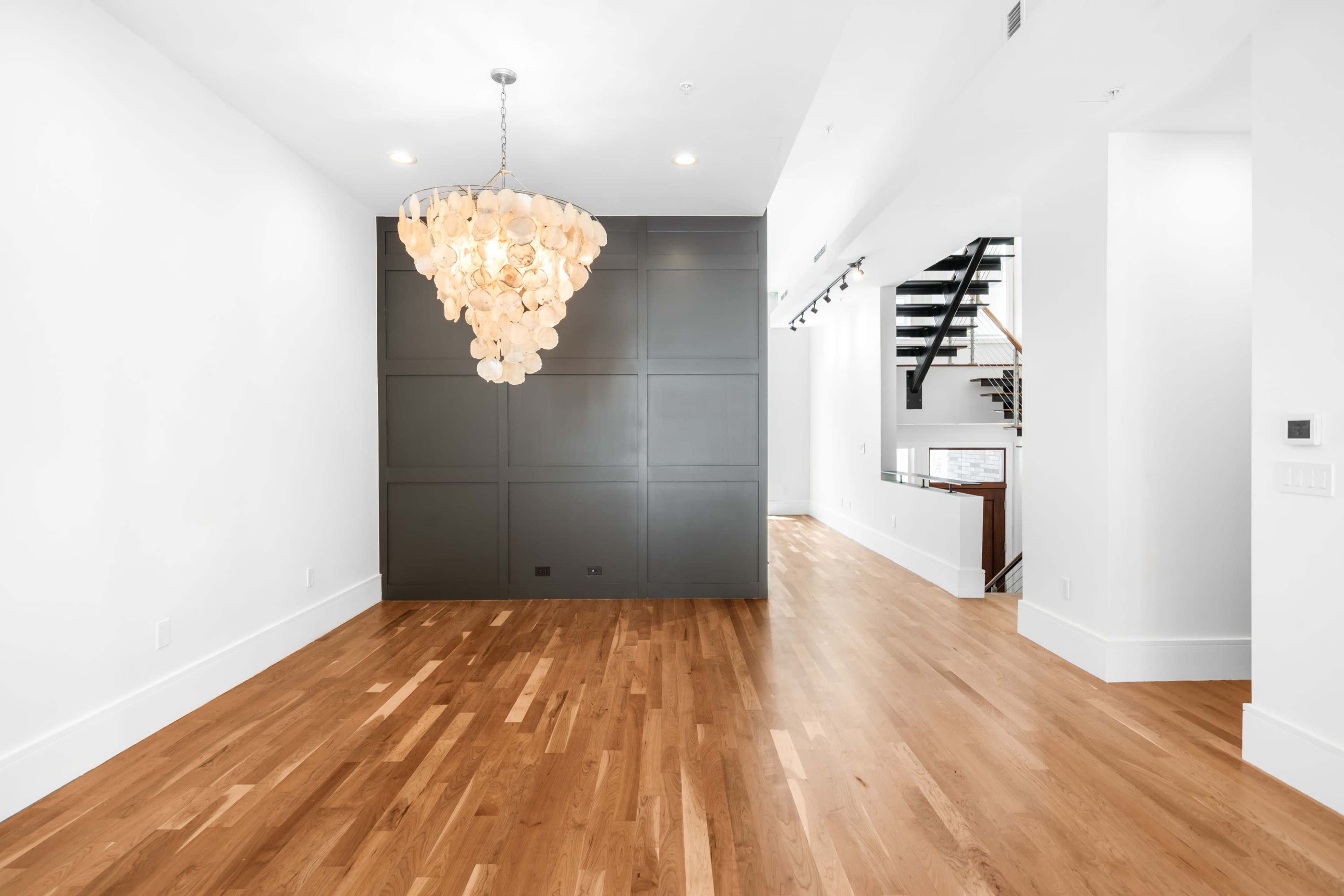 The image shows a spacious, modern interior featuring hardwood flooring, a large chandelier, and a staircase in the background.