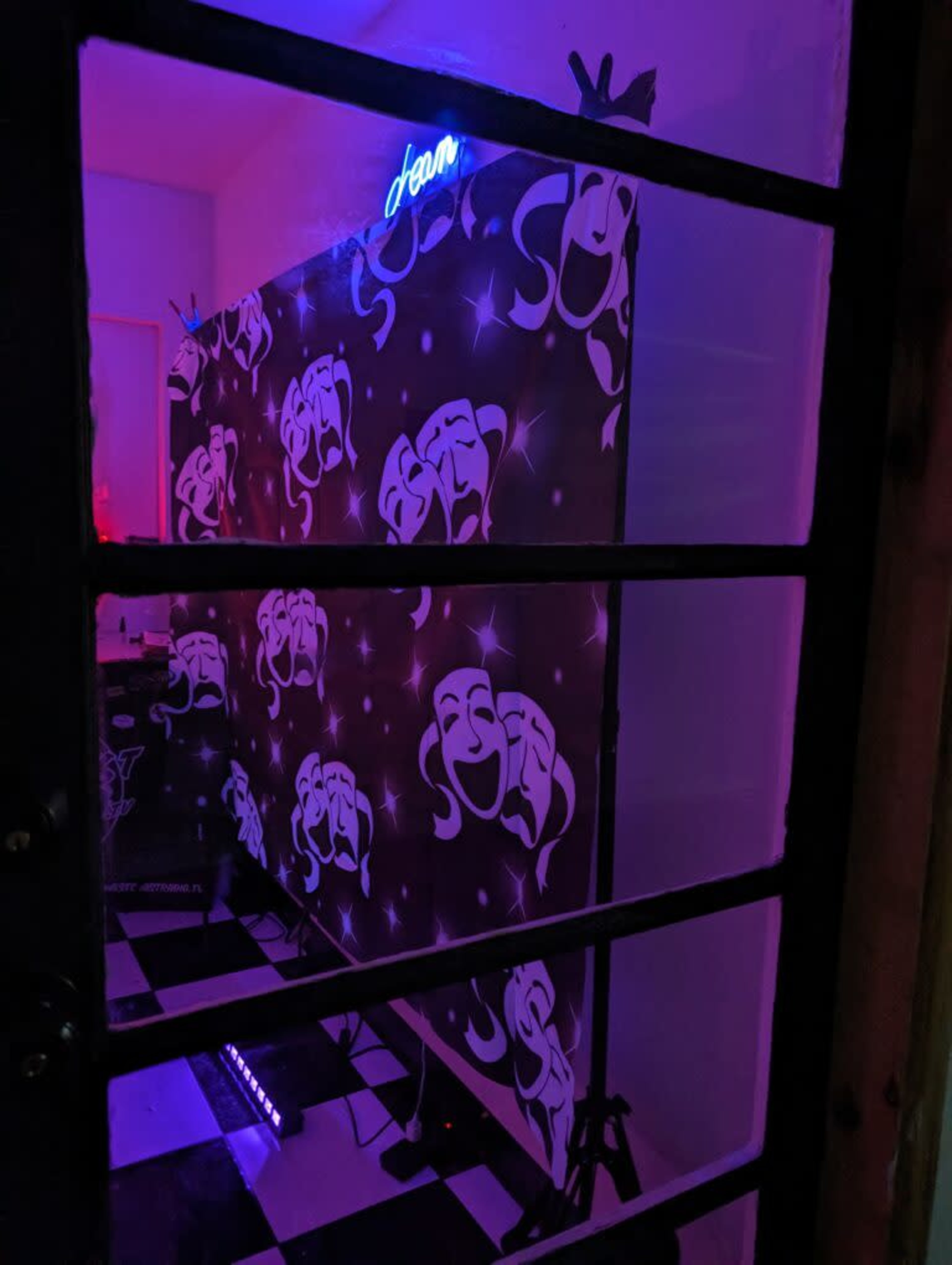 A room with a black and white checkered floor and a backdrop featuring a stylized design, illuminated by purple lighting and a neon sign that reads "Jean."