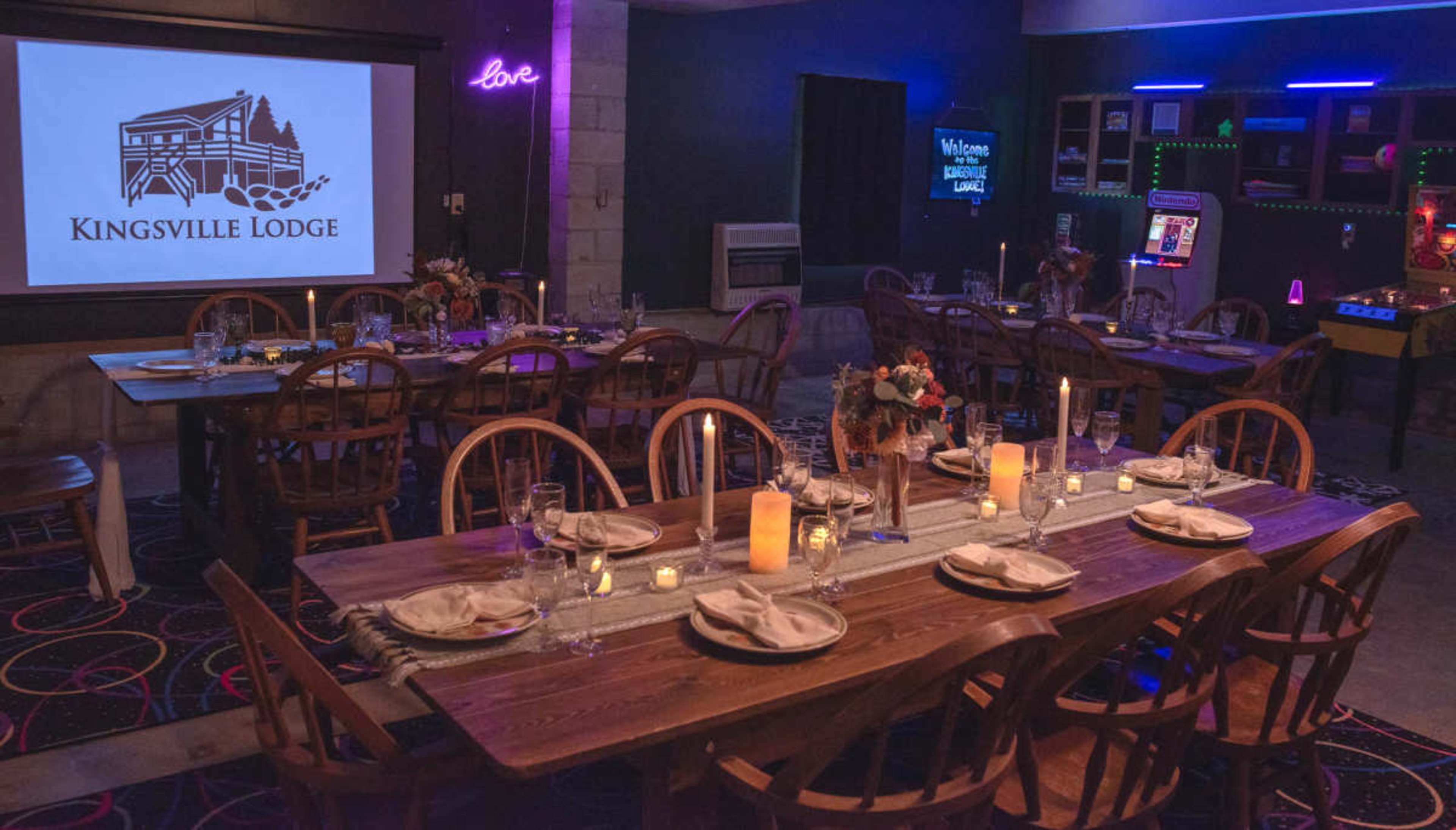The image shows a dining area with wooden tables set for a meal, featuring candles and flower centerpieces, alongside a logo of "Kingsville Lodge" displayed on a screen.
