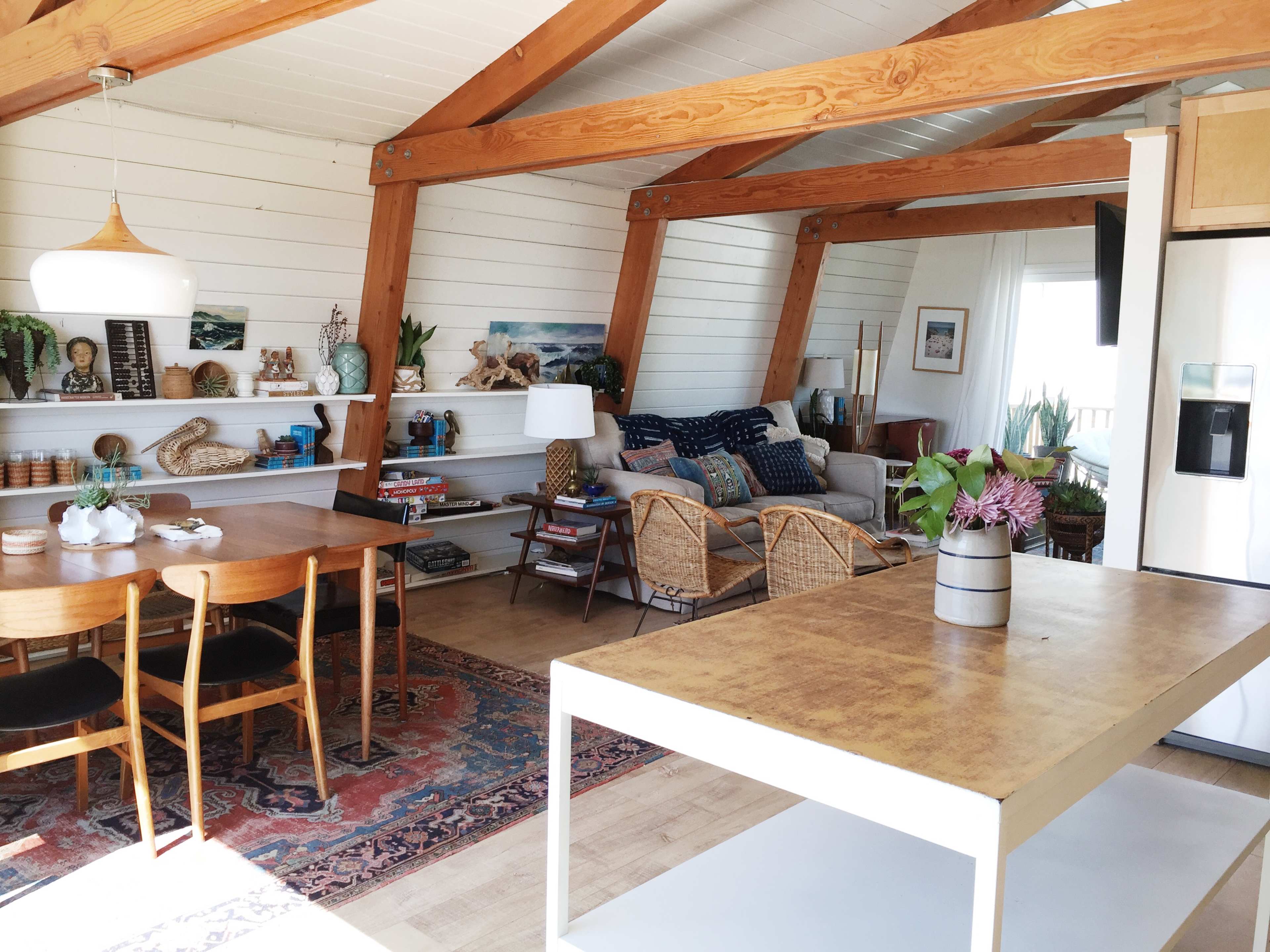 The image shows a bright and airy interior of a home with a dining table, a living area with a sofa and chairs, and wooden beams supporting the ceiling.