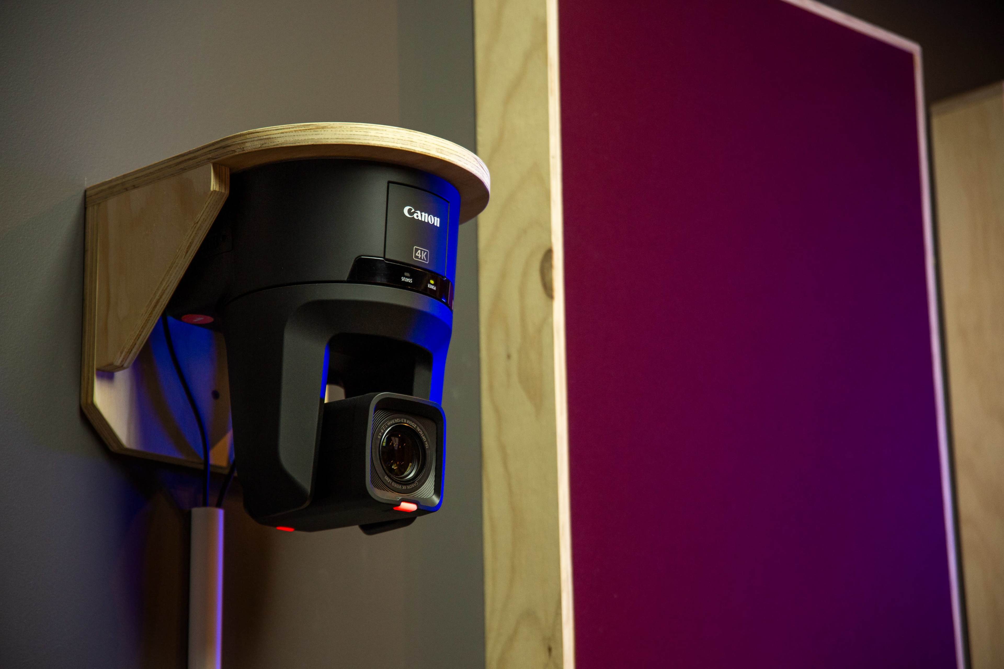 A Canon camera is mounted on a wooden shelf against a gray wall, with a purple-colored panel nearby.