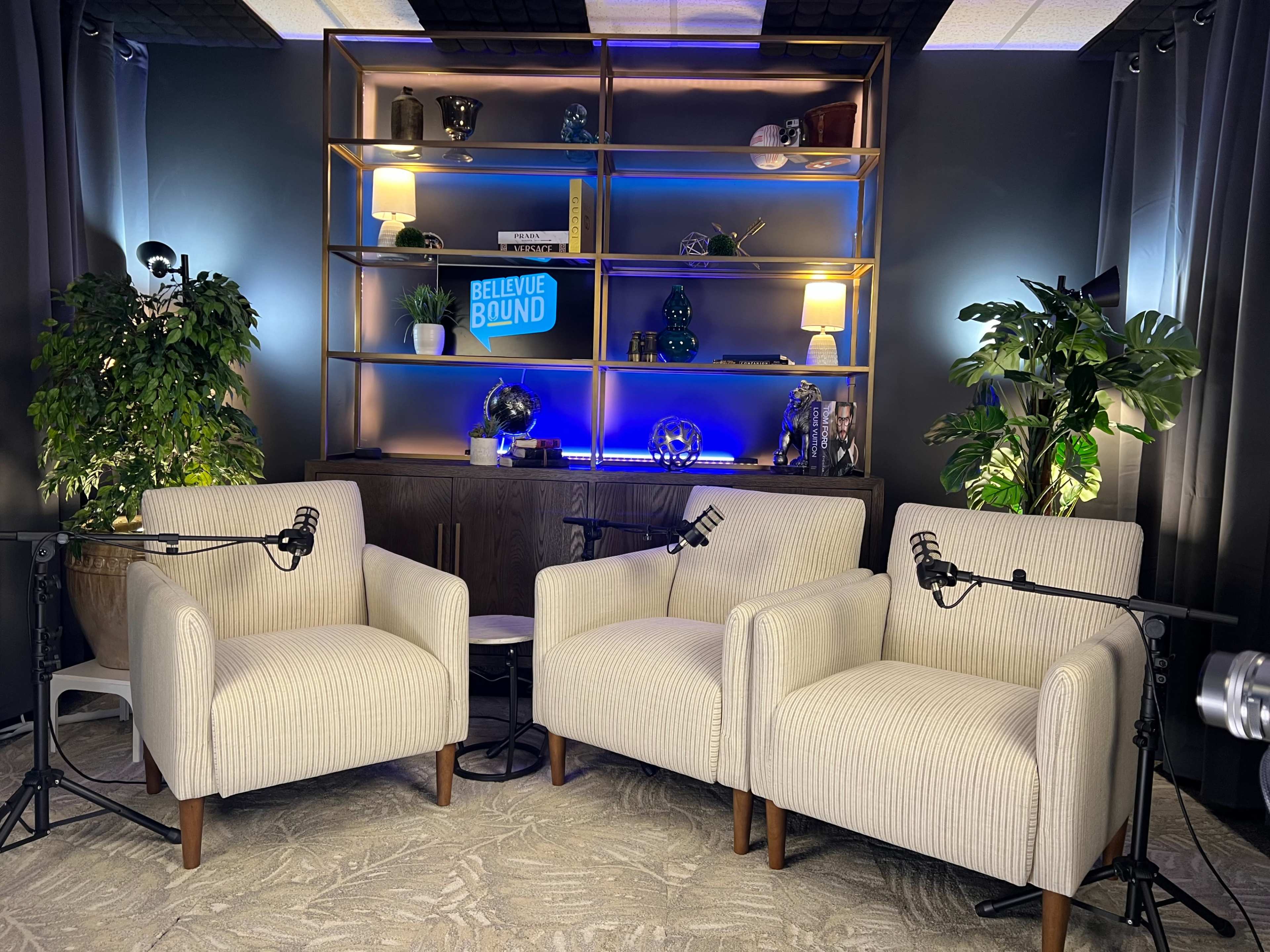 The image shows a cozy studio setup with two cream-colored armchairs, a wooden shelf with decorative items, and subtle lighting.