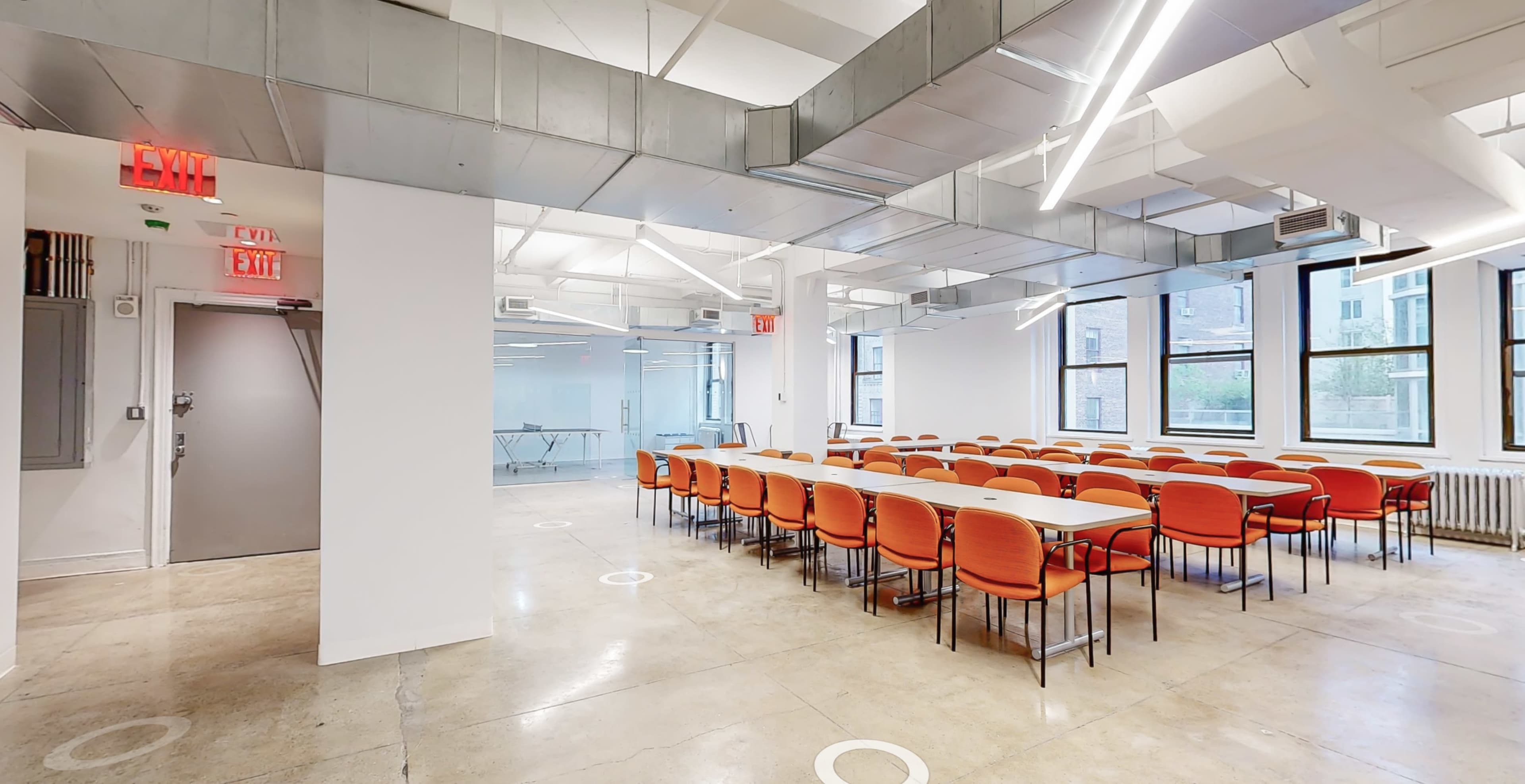 A spacious meeting room with rows of orange chairs and long tables arranged for a gathering, featuring large windows and an industrial ceiling design.