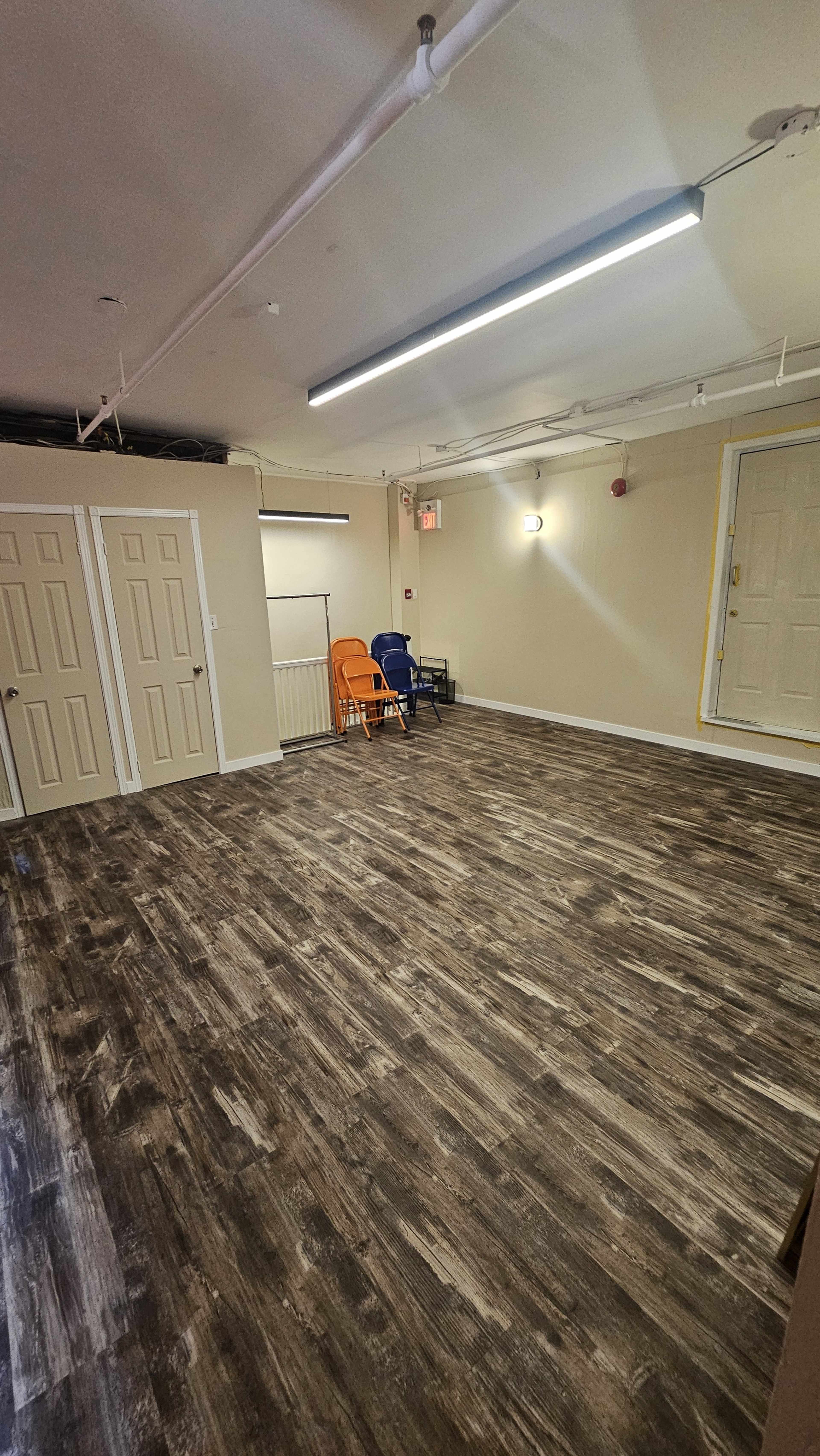 The image shows an empty room with a wooden floor, two chairs positioned against the wall, and several closed doors.