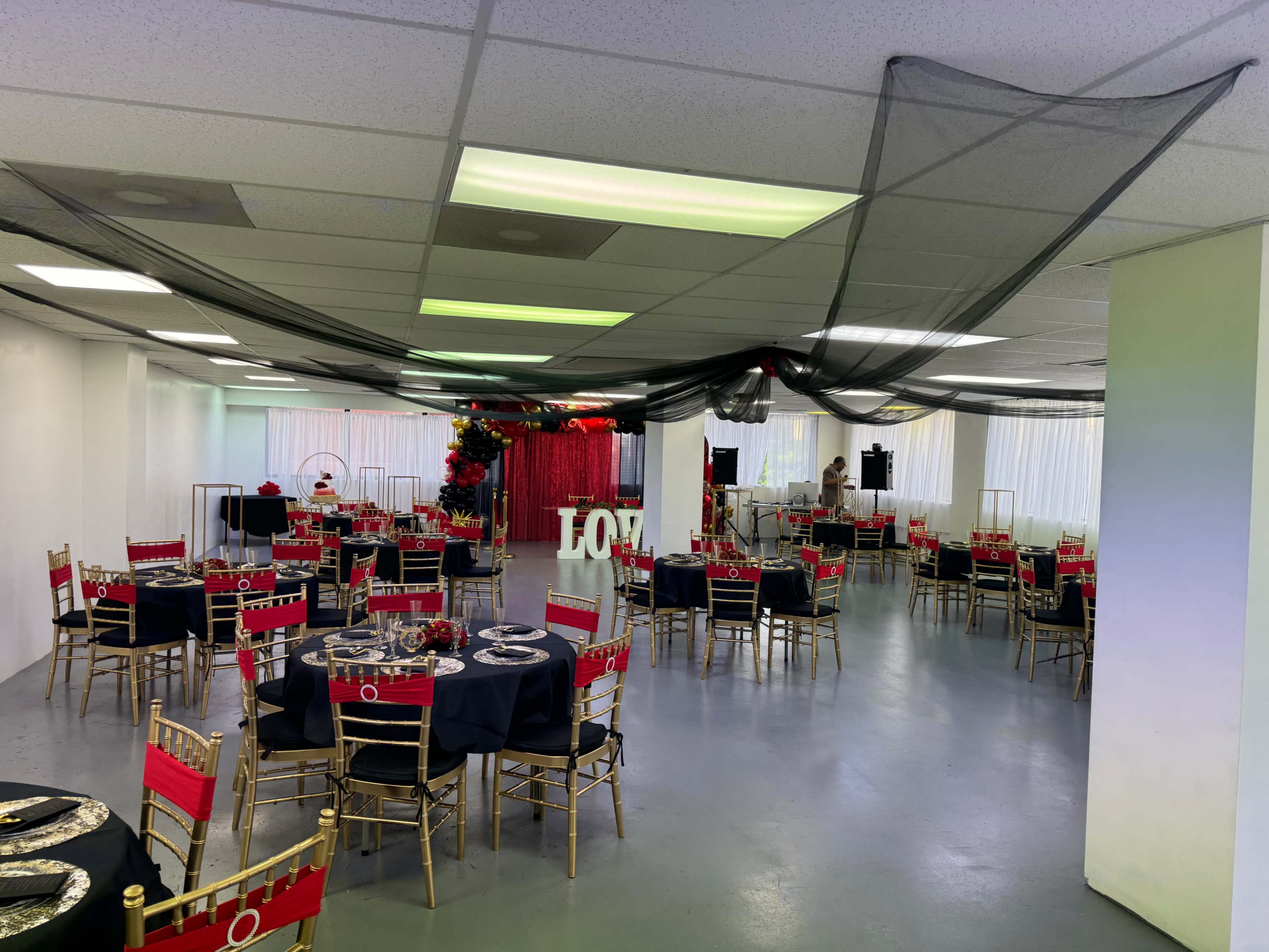 The image shows a spacious event venue set up for a formal gathering, featuring round tables with black tablecloths and red accents, gold chairs, and decorative elements like red drapes and floral arrangements.