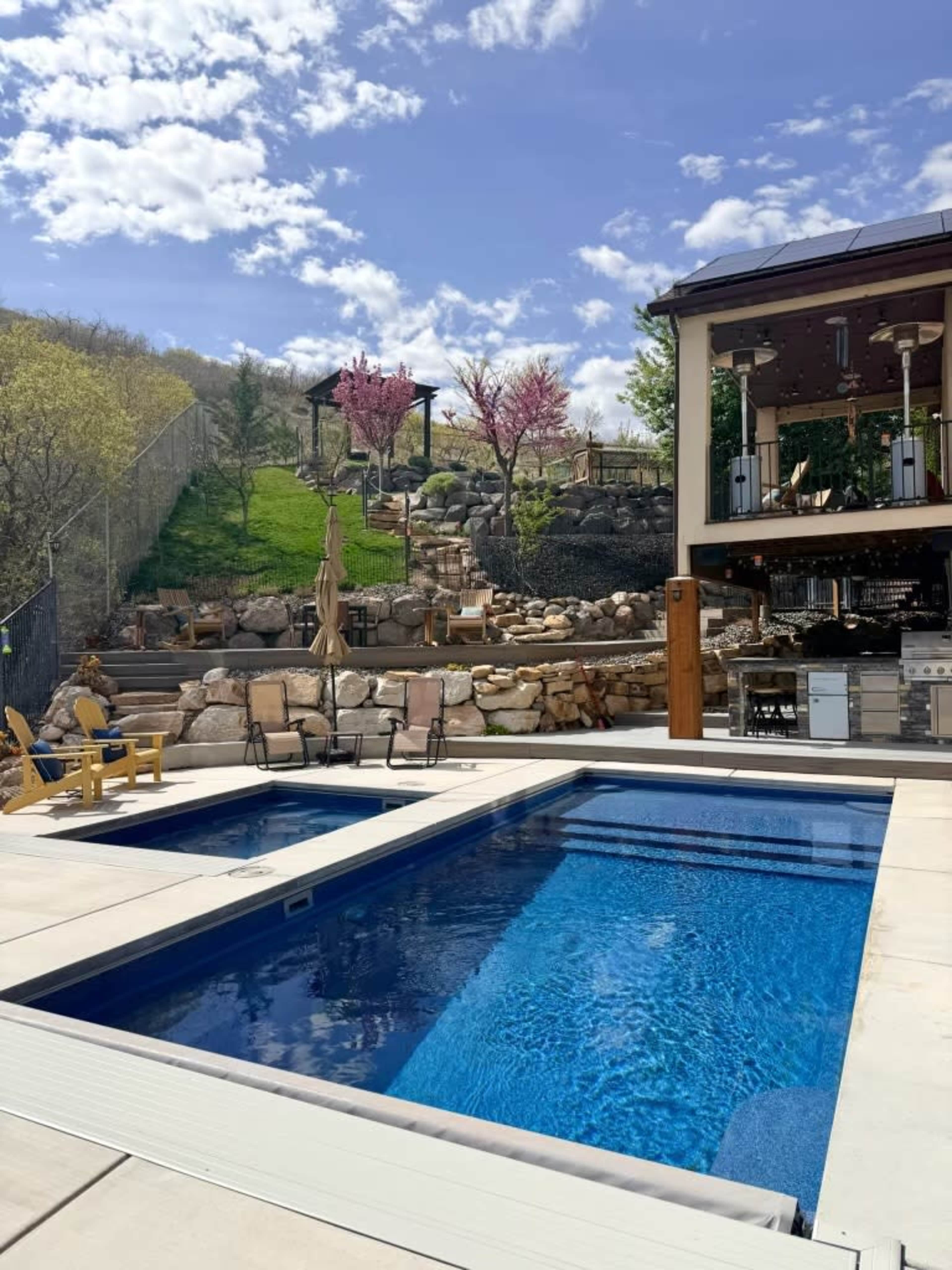 The image shows a swimming pool with a lounge area surrounded by stone landscaping and a slope featuring trees and outdoor seating in the background.