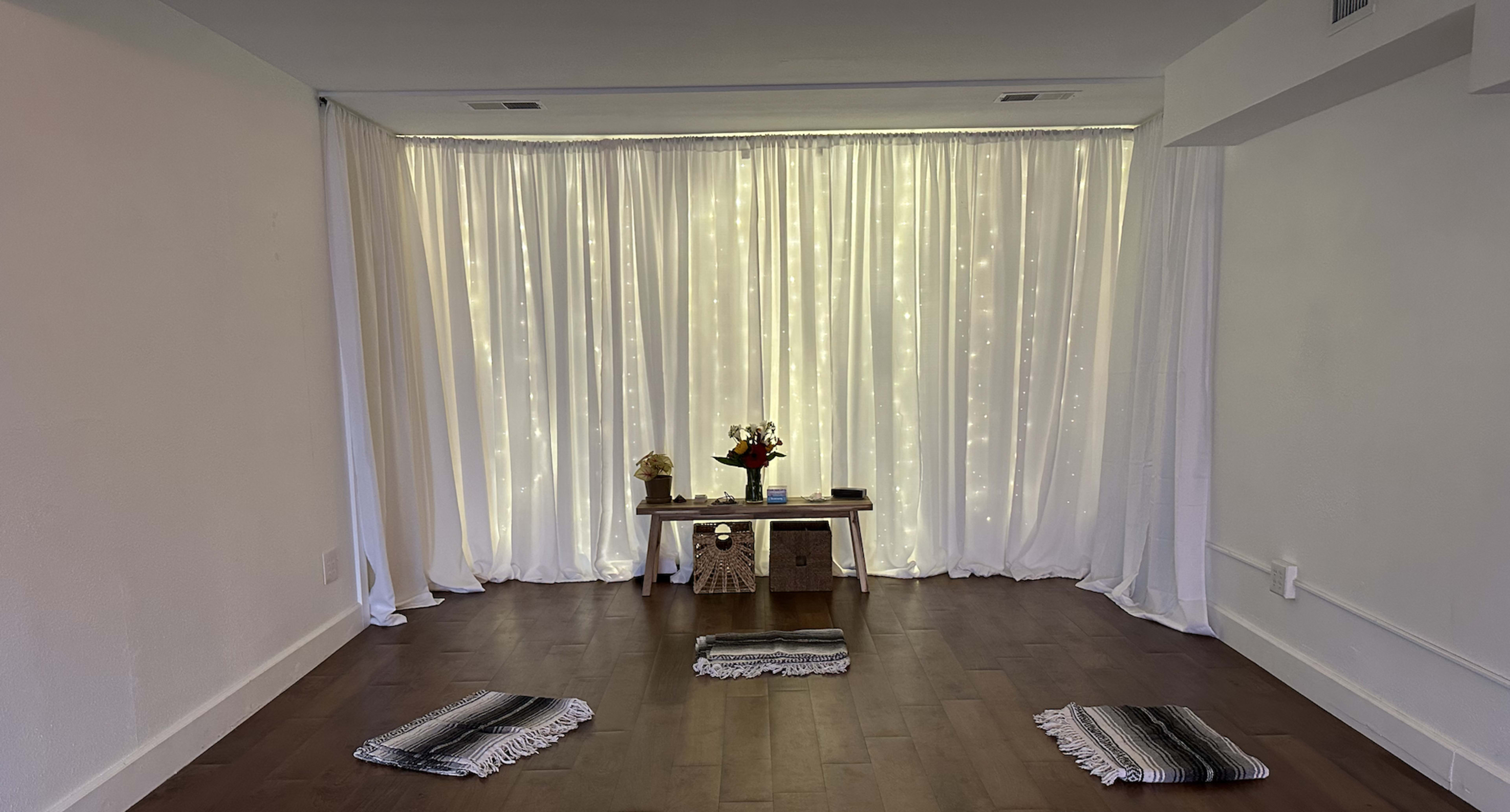 A room with a wooden table and a vase of flowers, surrounded by three small woven mats, and draped in white curtains with twinkling lights in the background.