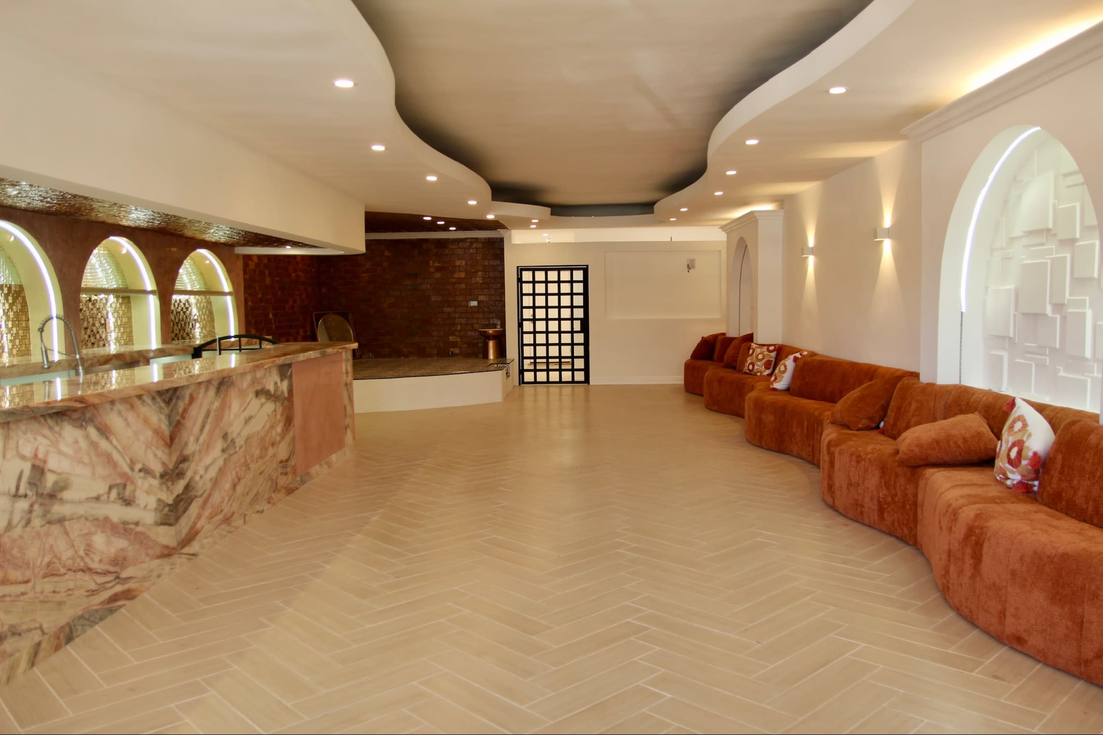 The image shows a spacious, modern interior featuring a curved sofa, a bar area with stone countertops, and decorative wall elements.