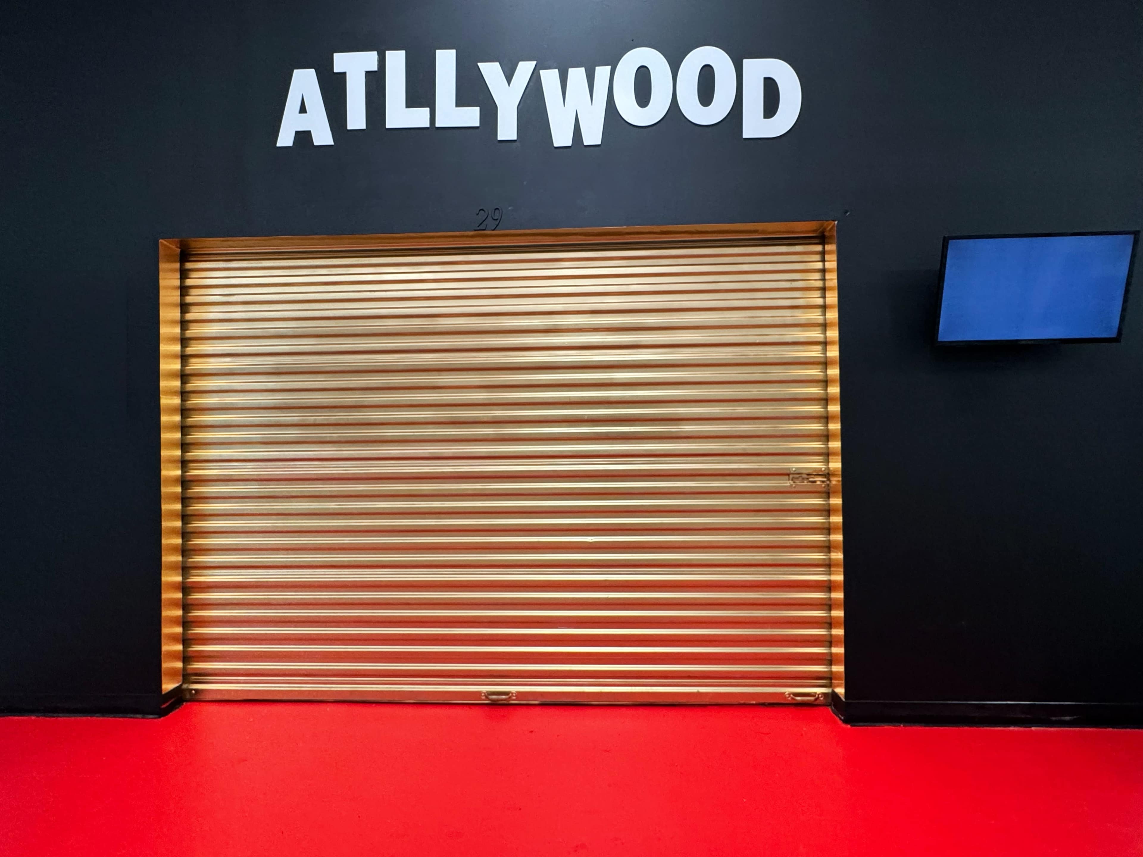 The image shows a large, closed golden roll-up door with the word "ATLLYWOOD" displayed above it on a black wall, against a red floor.