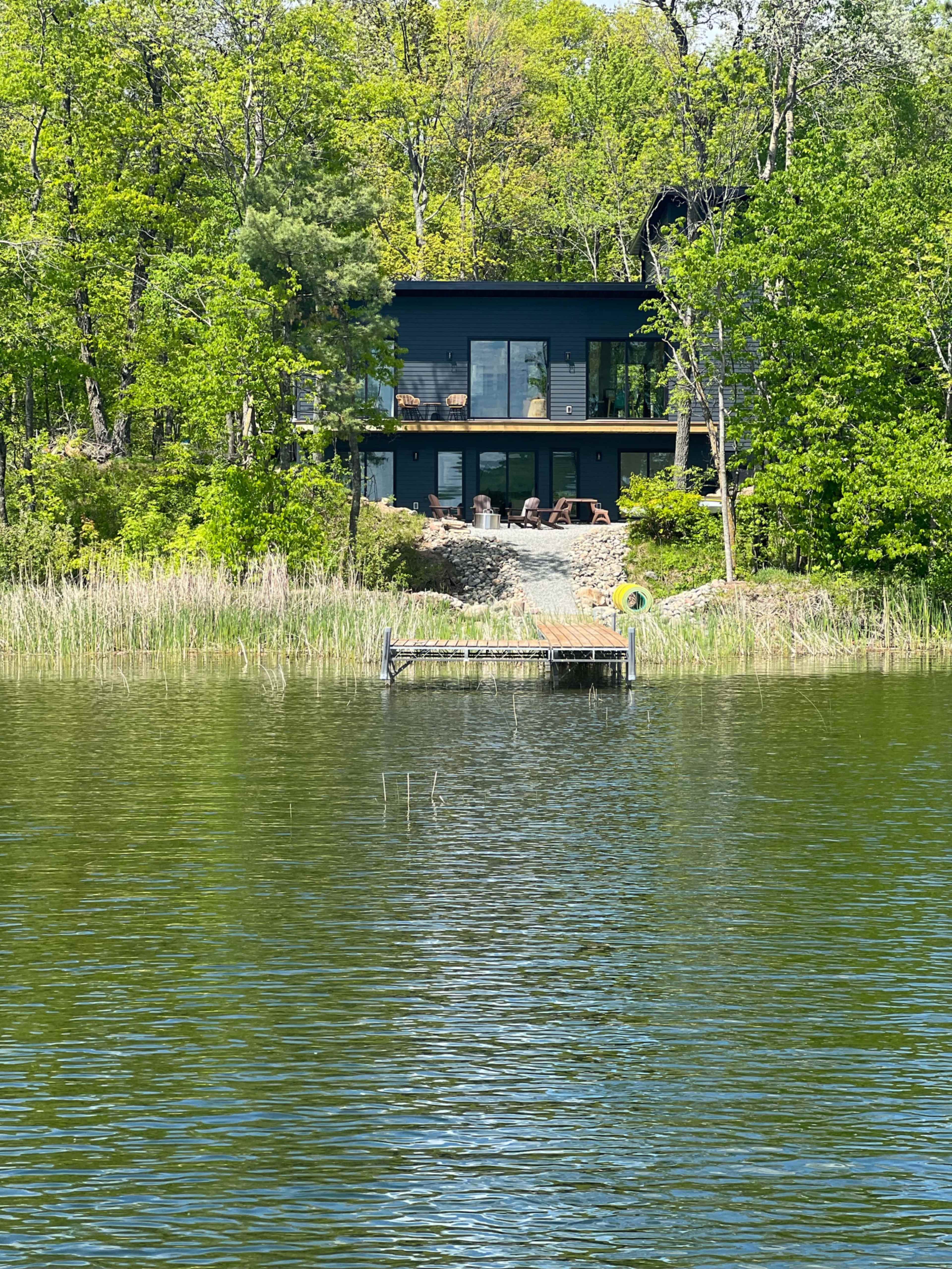 The image shows a modern black house overlooking a calm lake, surrounded by lush greenery and a wooden dock extending into the water.