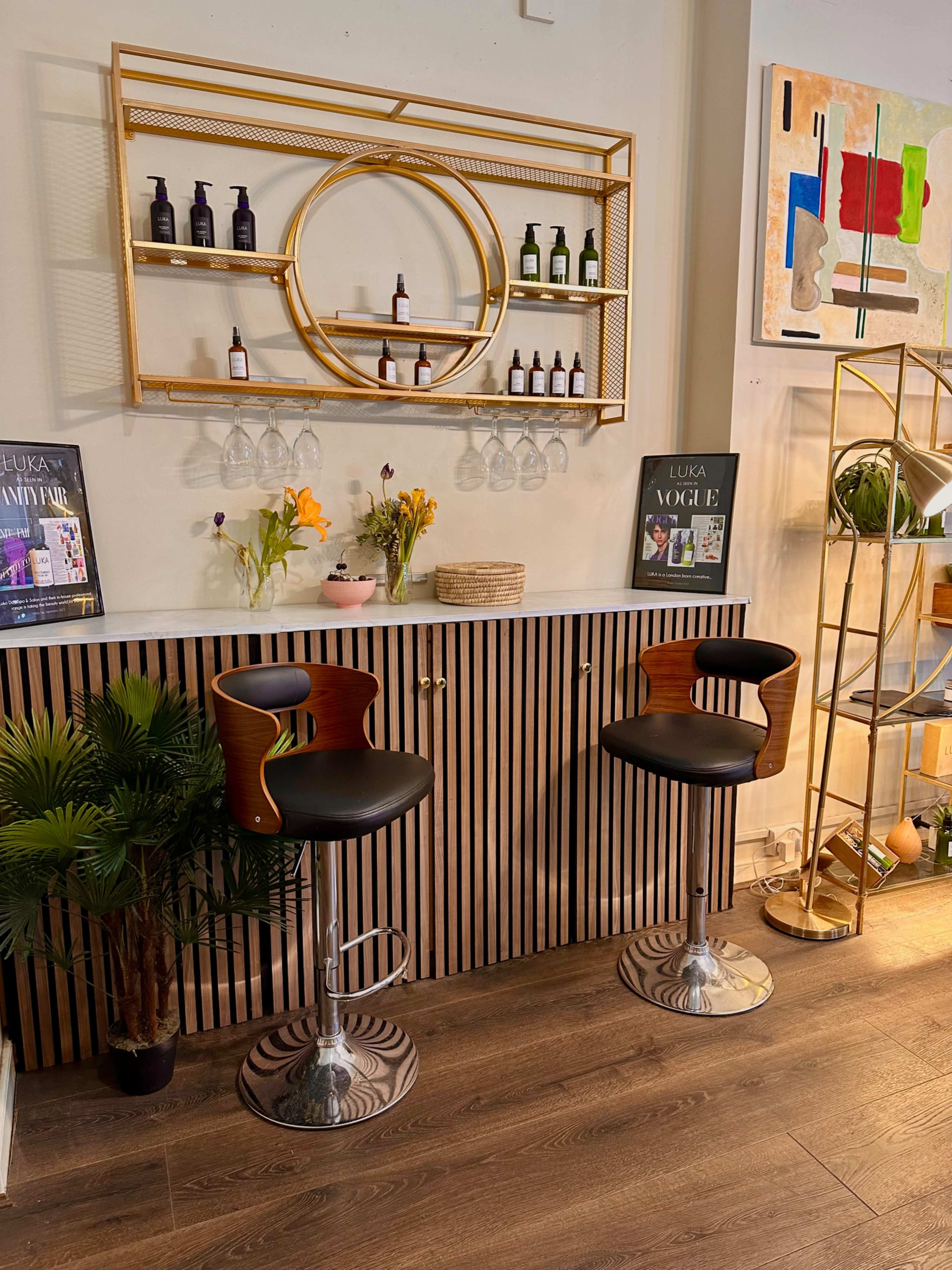 The image shows a modern interior featuring two black bar stools at a counter, with a gold circular shelf above displaying various bottles and decorative items.