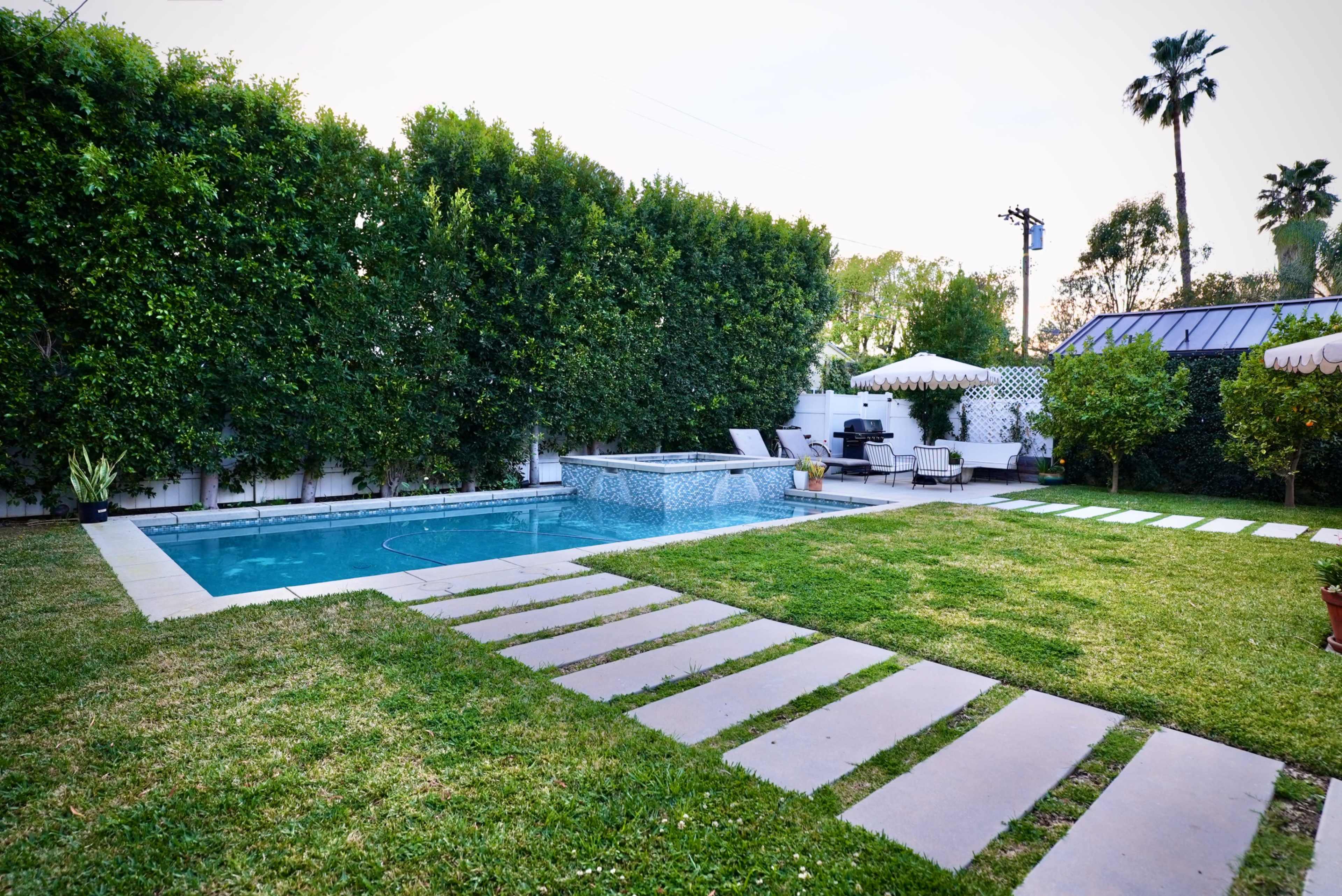 The image shows a backyard with a swimming pool, stone pathway, and well-maintained lawn surrounded by tall hedges.