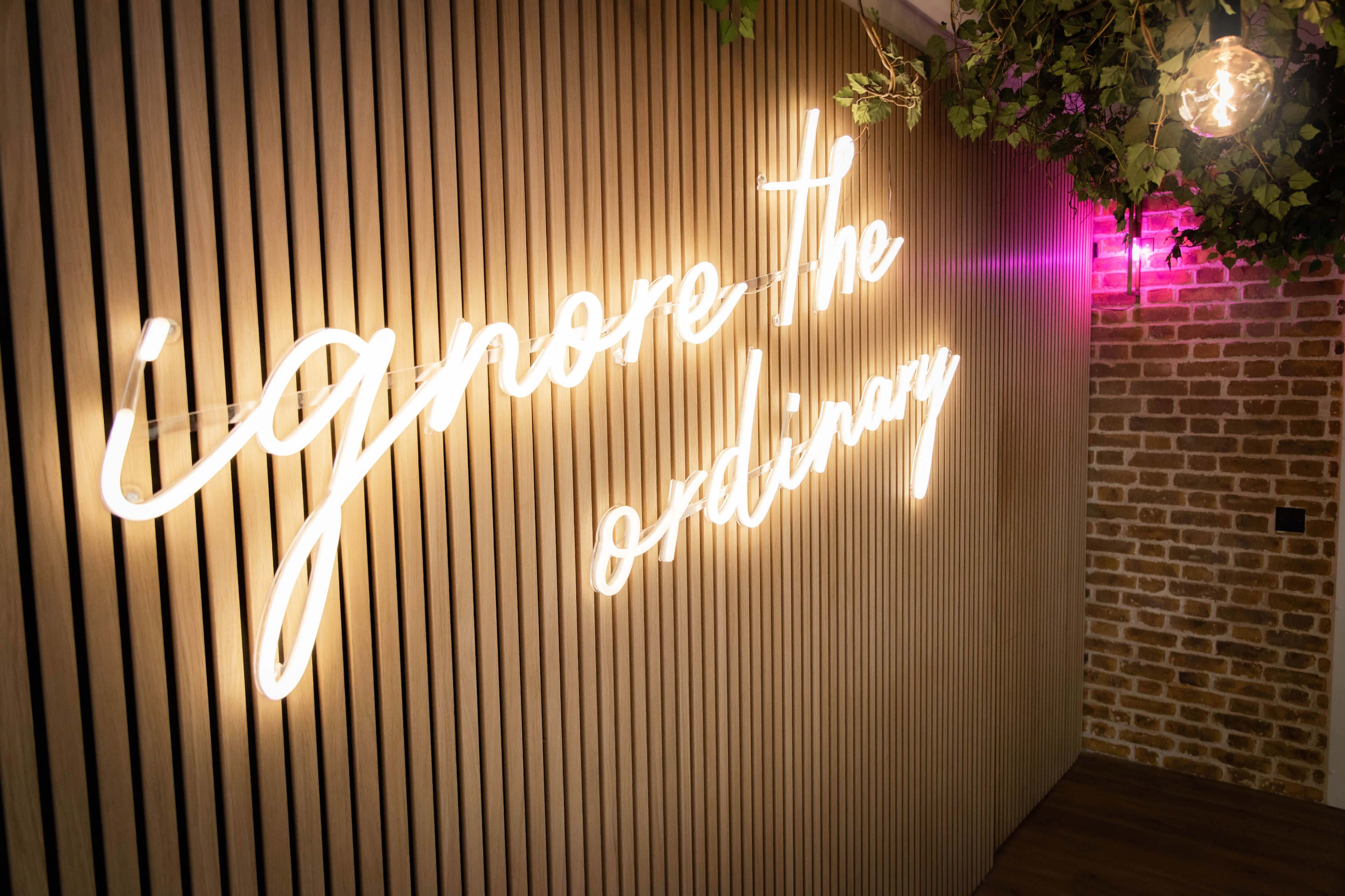 A neon sign that reads "ignore the ordinary" is illuminated on a wooden slat wall with greenery overhead.