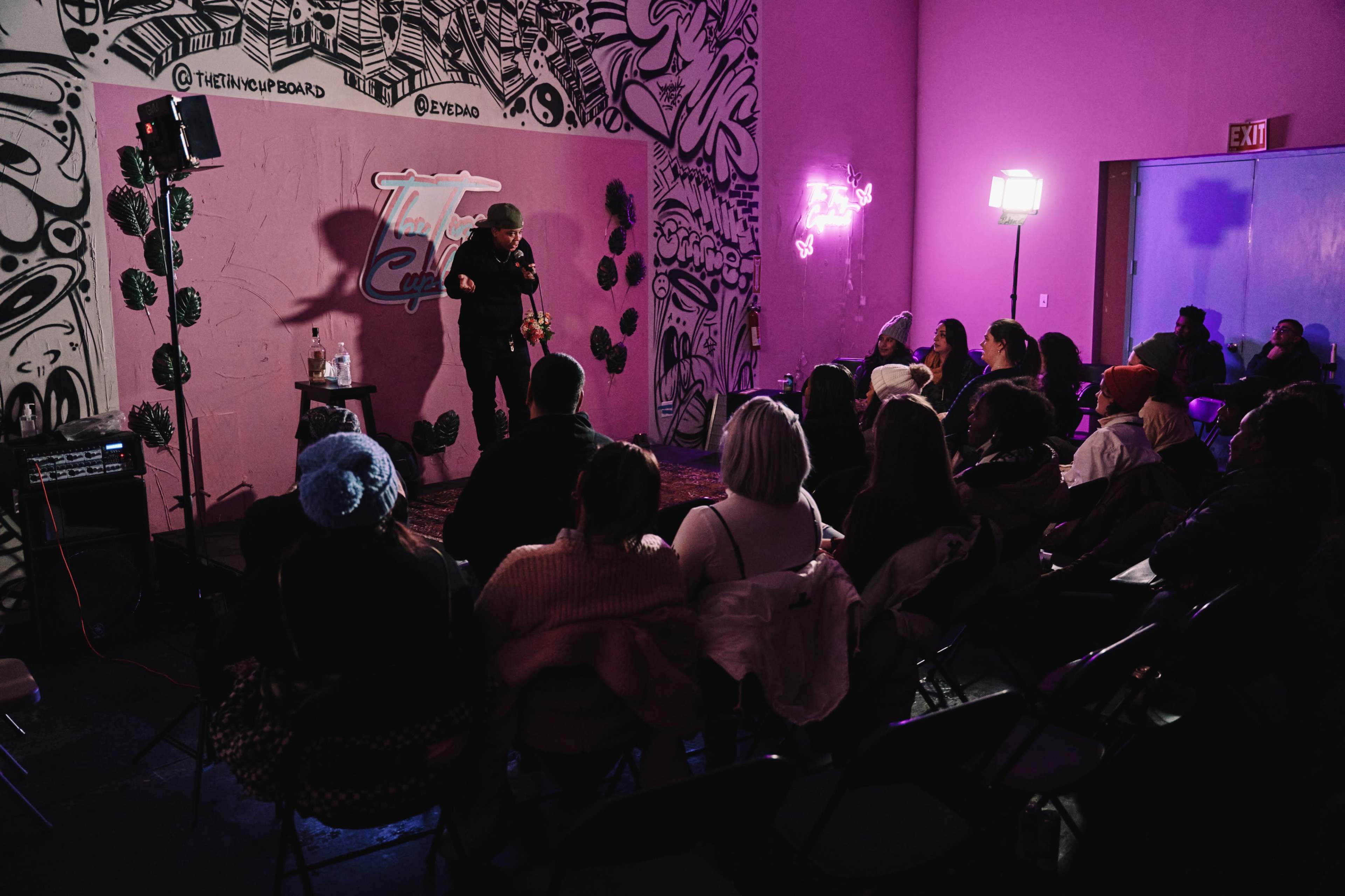 A performer stands on stage in front of an audience in a pink room adorned with graffiti and decorative lights.