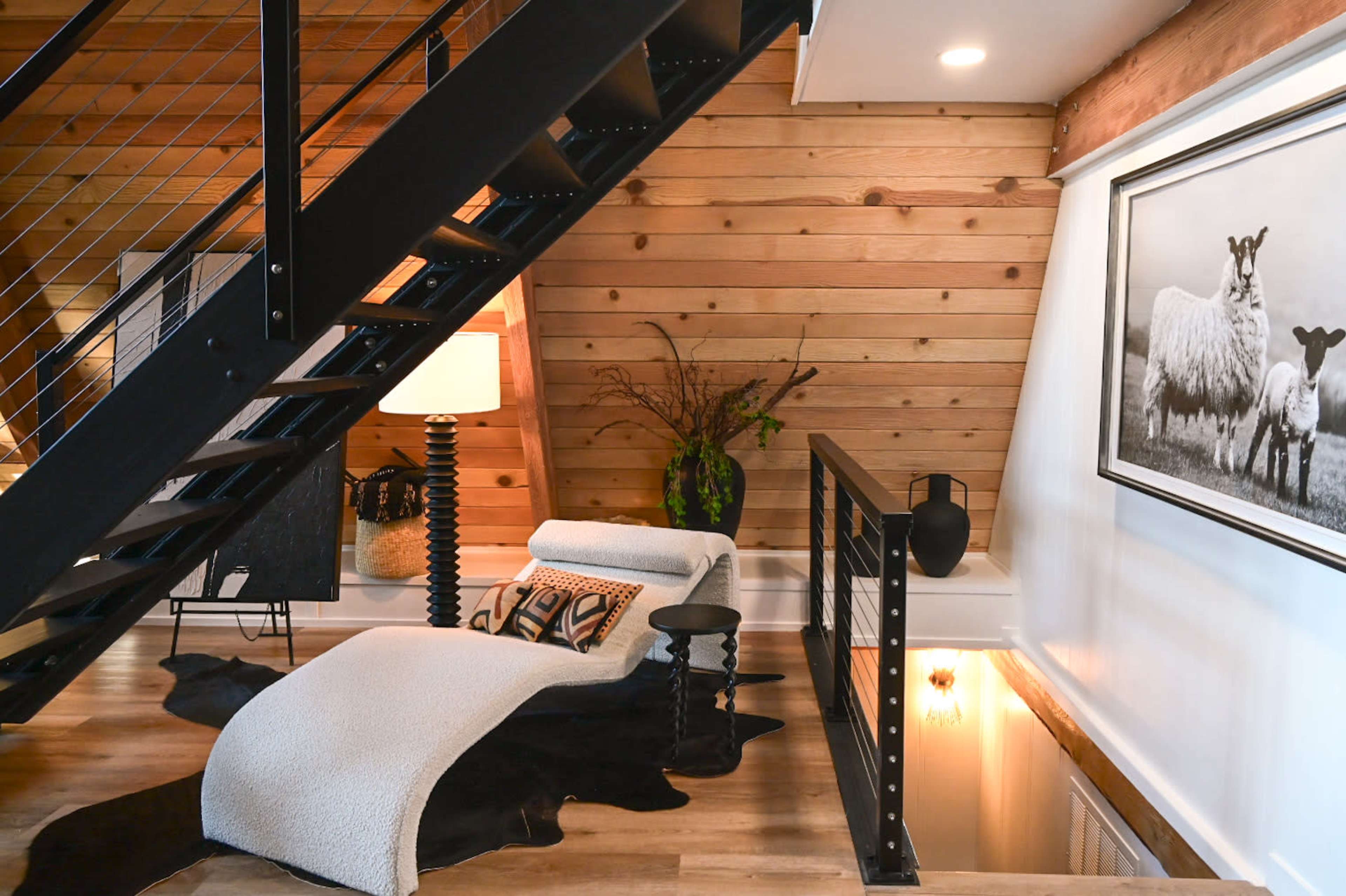 A staircase with a black metal railing leads to a cozy loft area featuring a chaise lounge, a lamp, and decorative elements against a wooden wall.