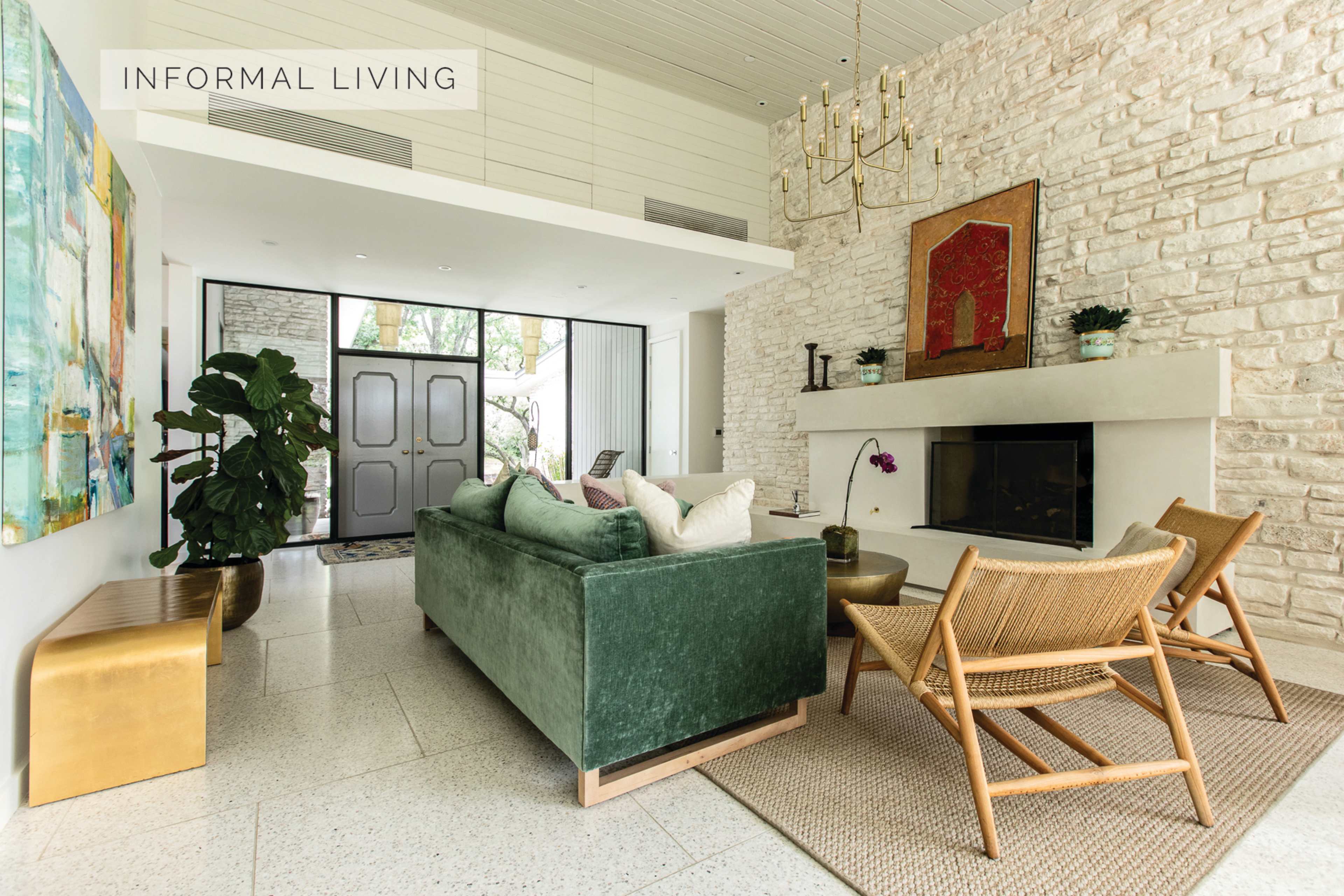 A modern living room with a stone wall, a fireplace, a green velvet sofa, and wooden chairs, alongside large glass doors.