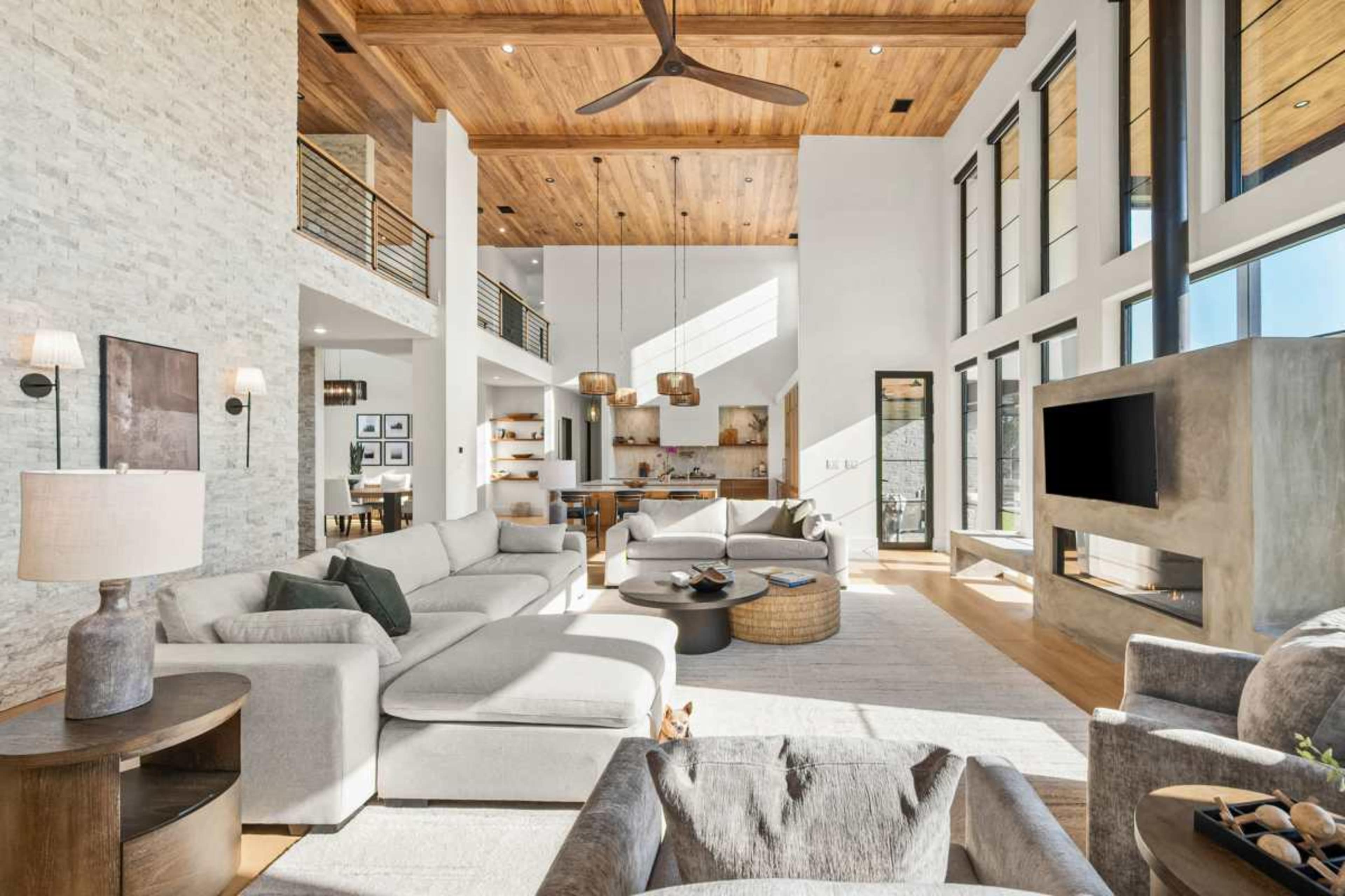 A spacious living room features large windows, a wooden ceiling, and modern furniture, creating a bright and inviting atmosphere.