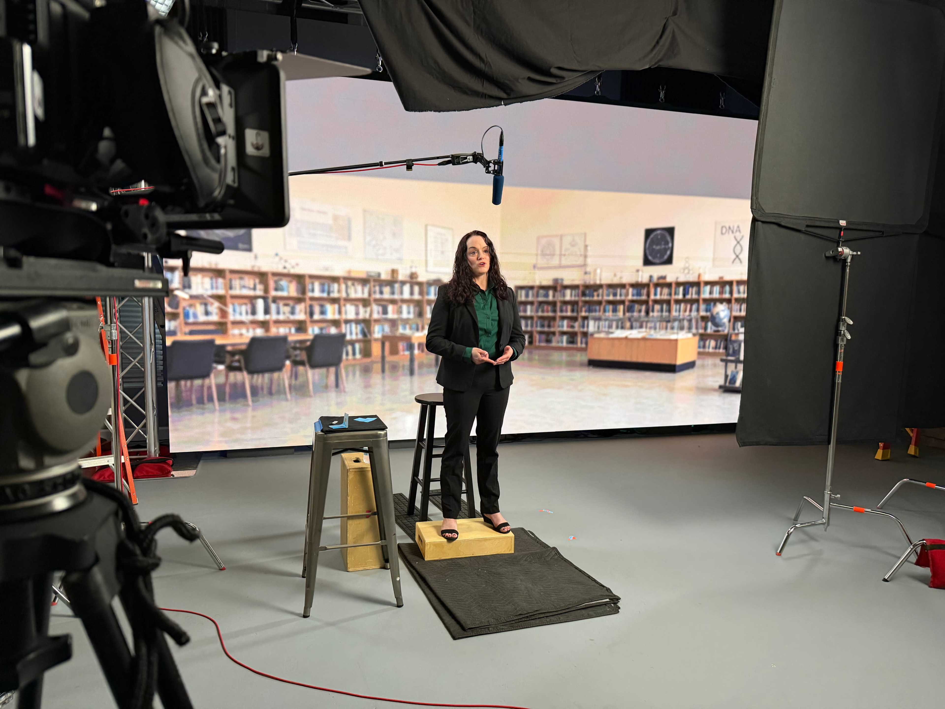 A woman stands on a small platform in a studio setting, presenting in front of a large backdrop that depicts a library.