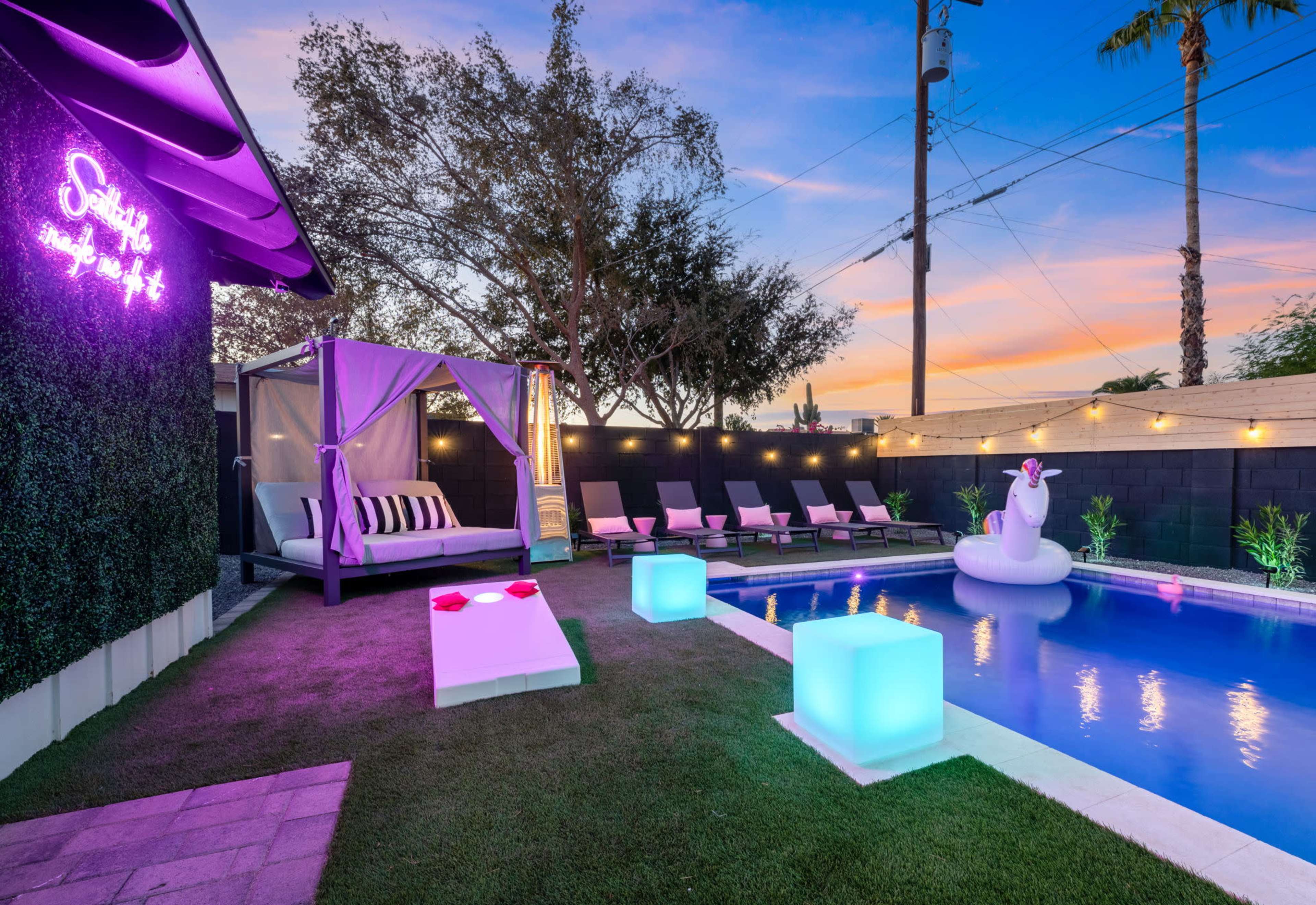 A backyard pool area featuring a lounge with a canopy, several deck chairs, illuminated decorative cubes, and a large unicorn float in the water.