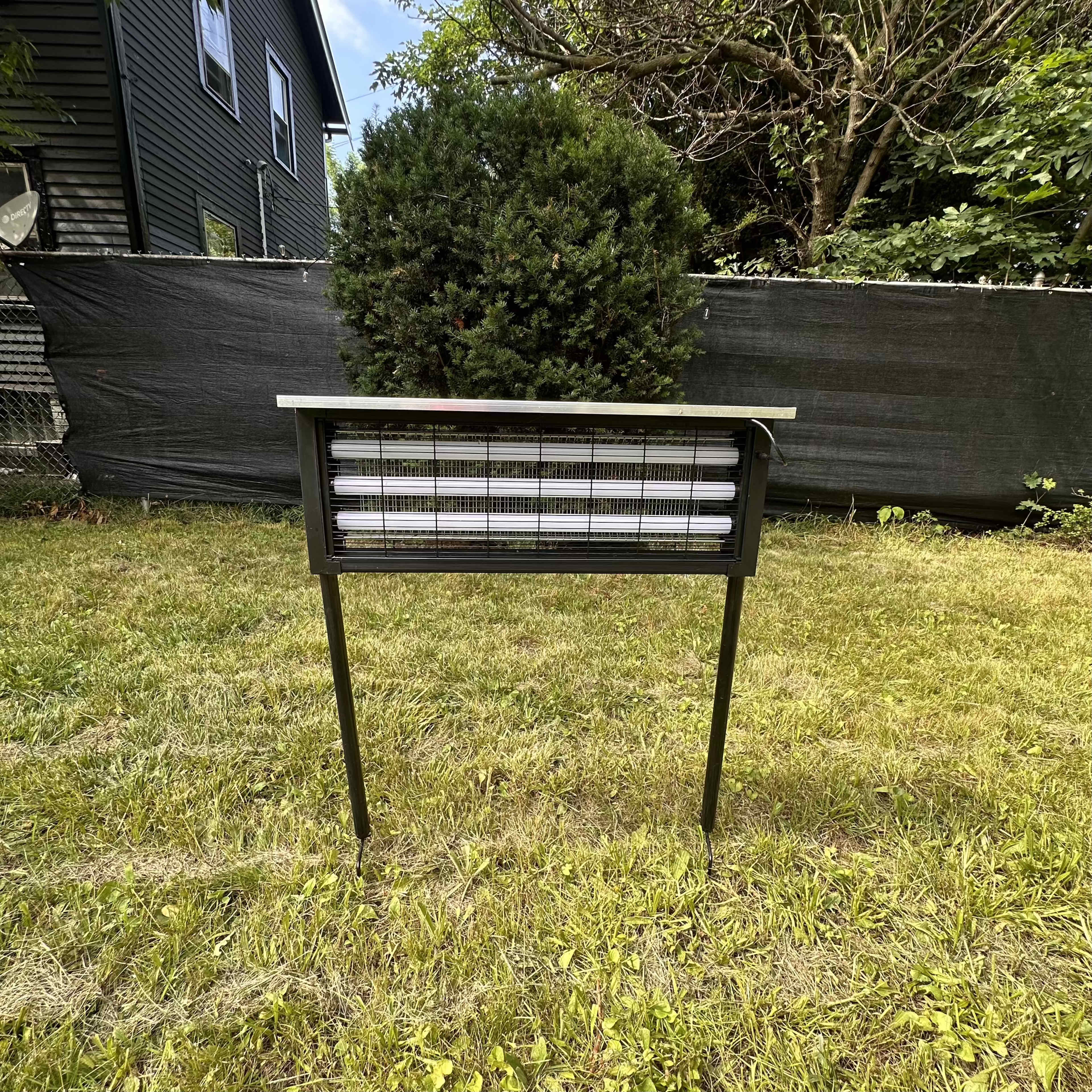 A standing, metallic insect zapper is positioned on a grassy area with a hedge and a dark fence in the background.