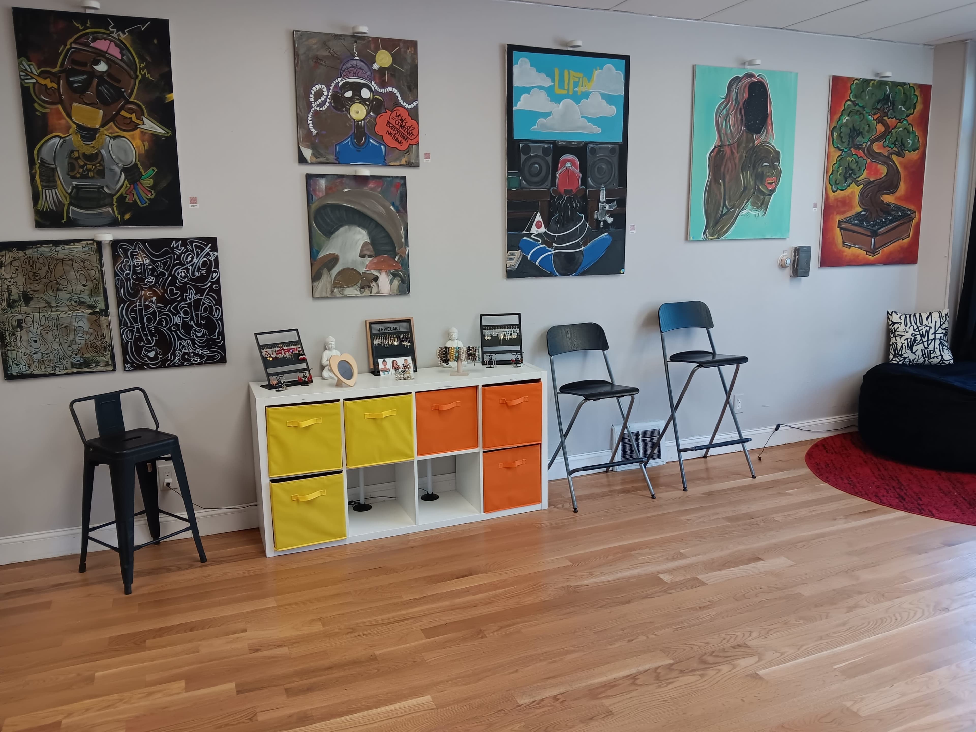 The image shows a gallery or studio space with colorful paintings on the walls, a white storage unit with yellow and orange drawers, and two black folding chairs arranged next to it.