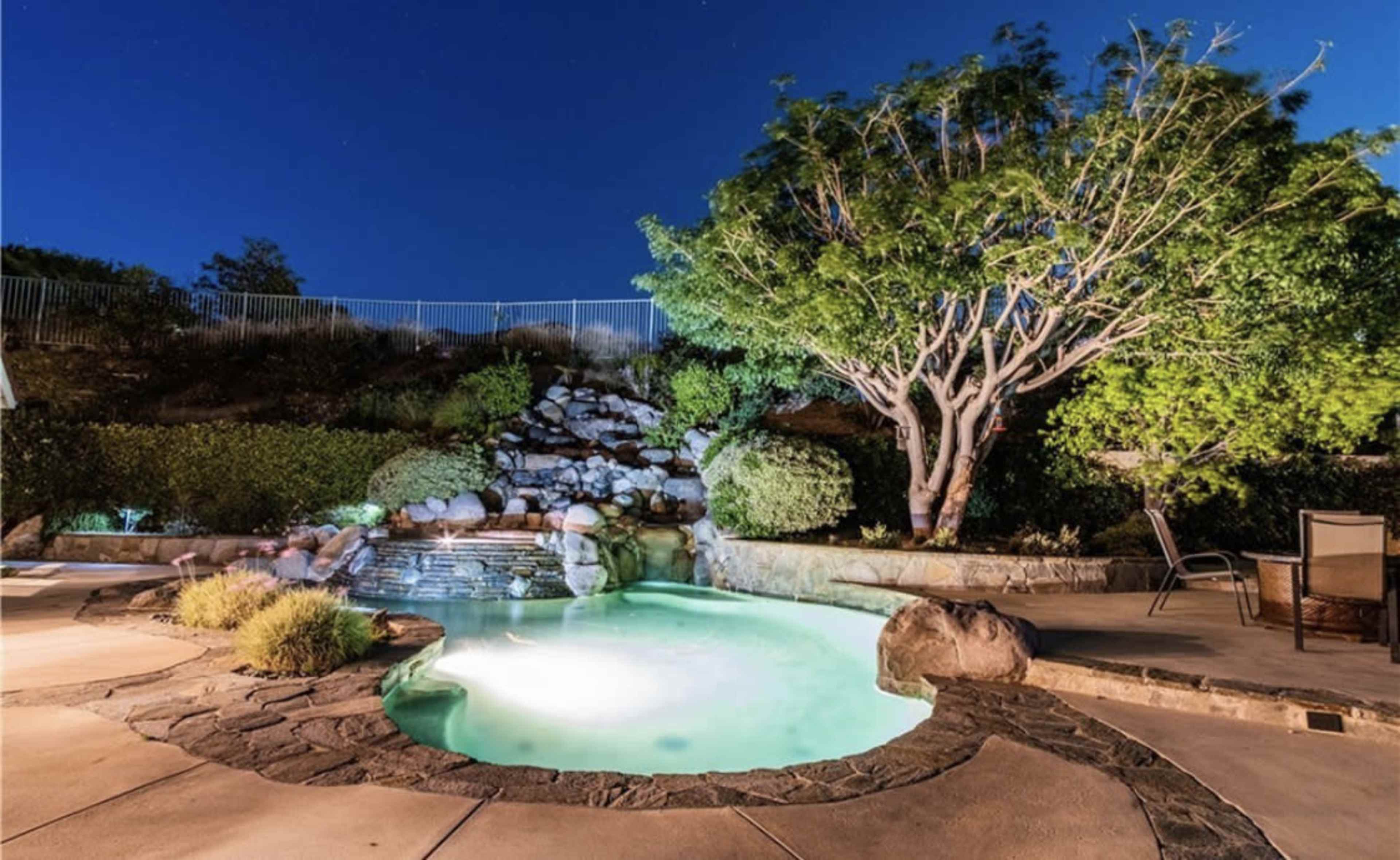 A backyard pool with a waterfall and landscaping is illuminated at night under a clear sky.