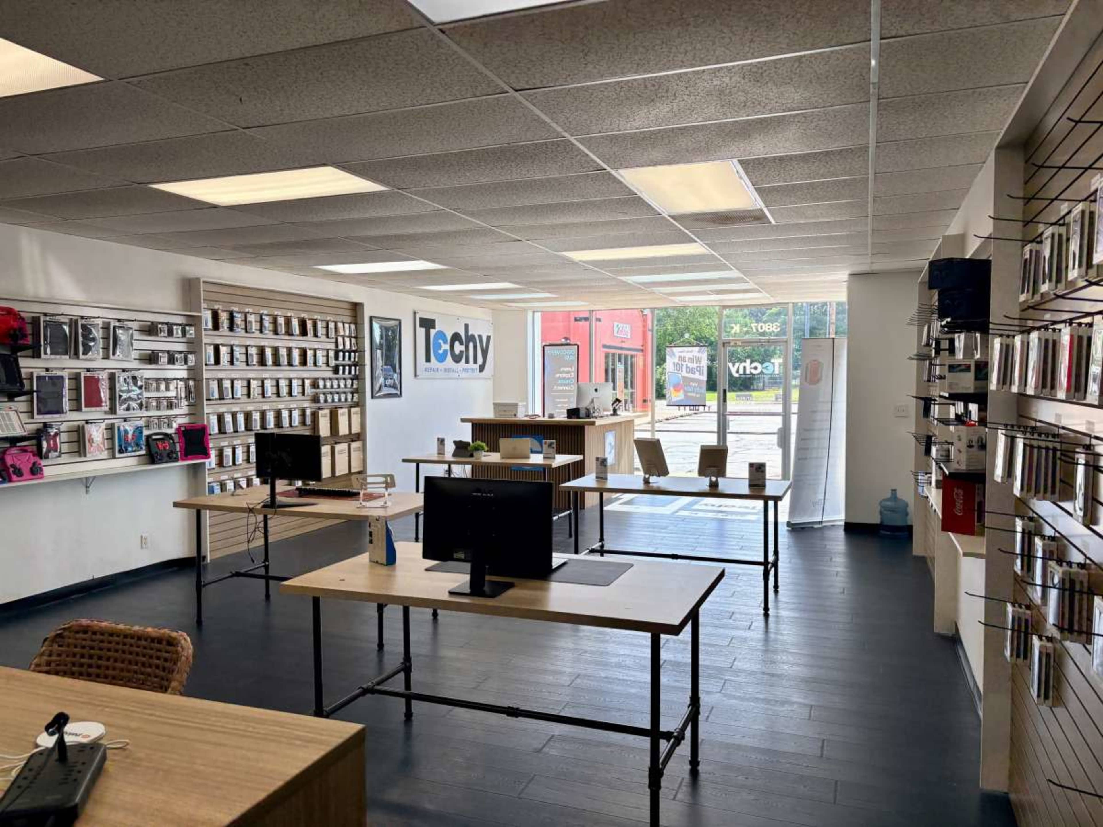 The image shows a retail store interior featuring neatly organized shelves of tech accessories and two tables with a computer setup.
