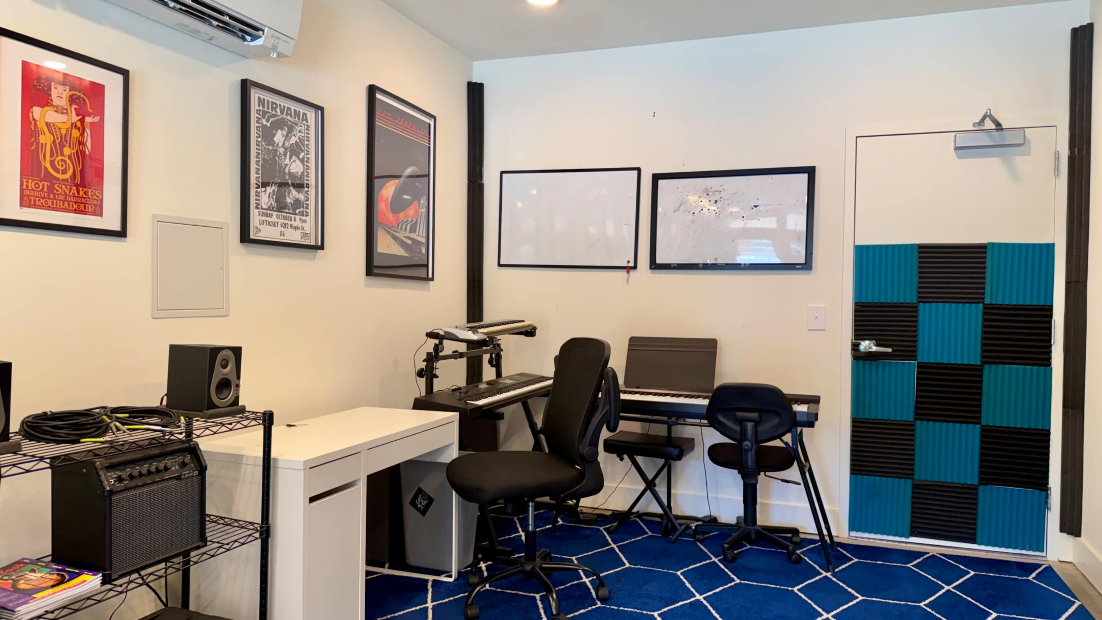 The image shows a small music studio featuring a keyboard, a desk with speakers, framed posters on the walls, and a blue-and-black accent wall by the door.