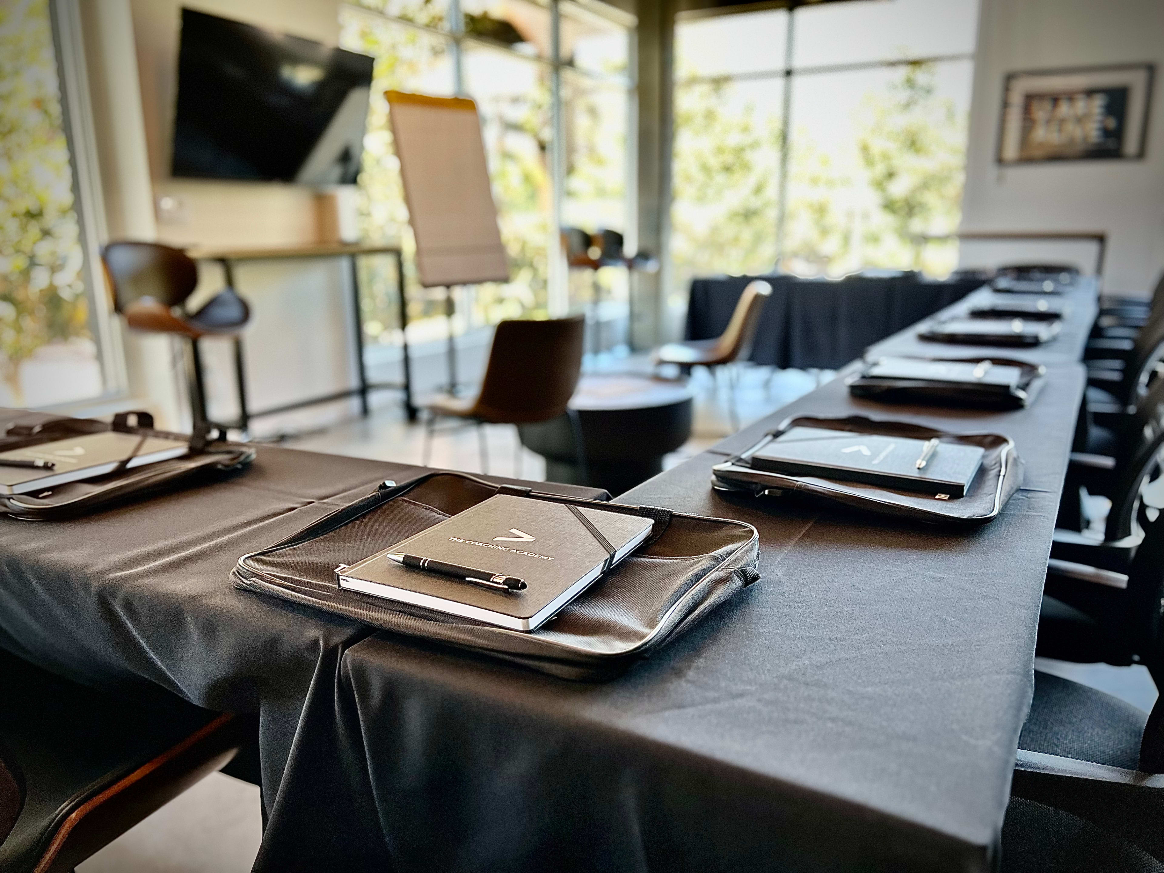 A boardroom is set up with a long table covered in black cloth, featuring notepads and pens on each place setting, alongside chairs and a flip chart in the background.