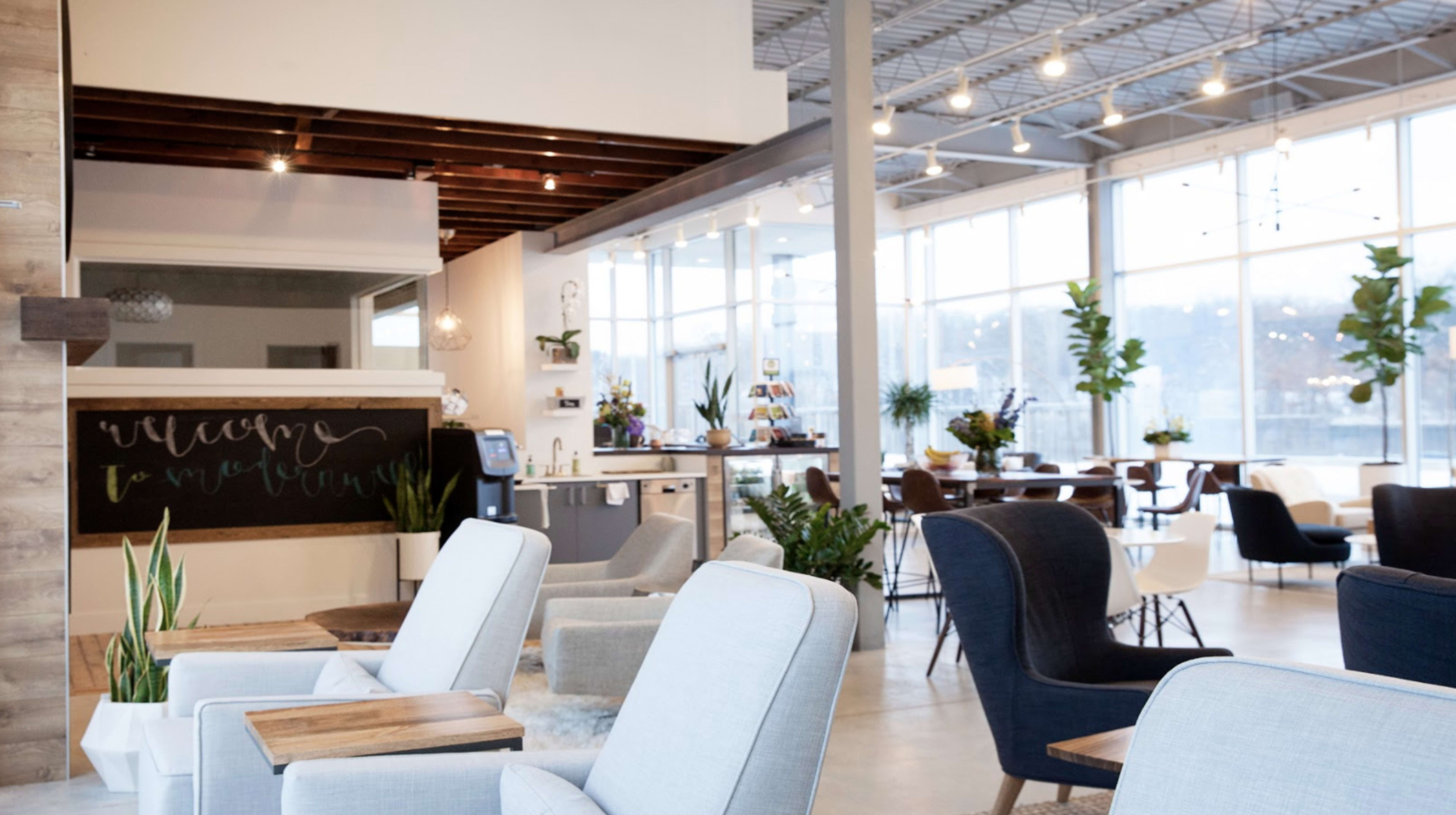 The image depicts a well-lit, modern lounge area featuring comfortable seating, plants, and a small café setup with a greeting message on the wall.