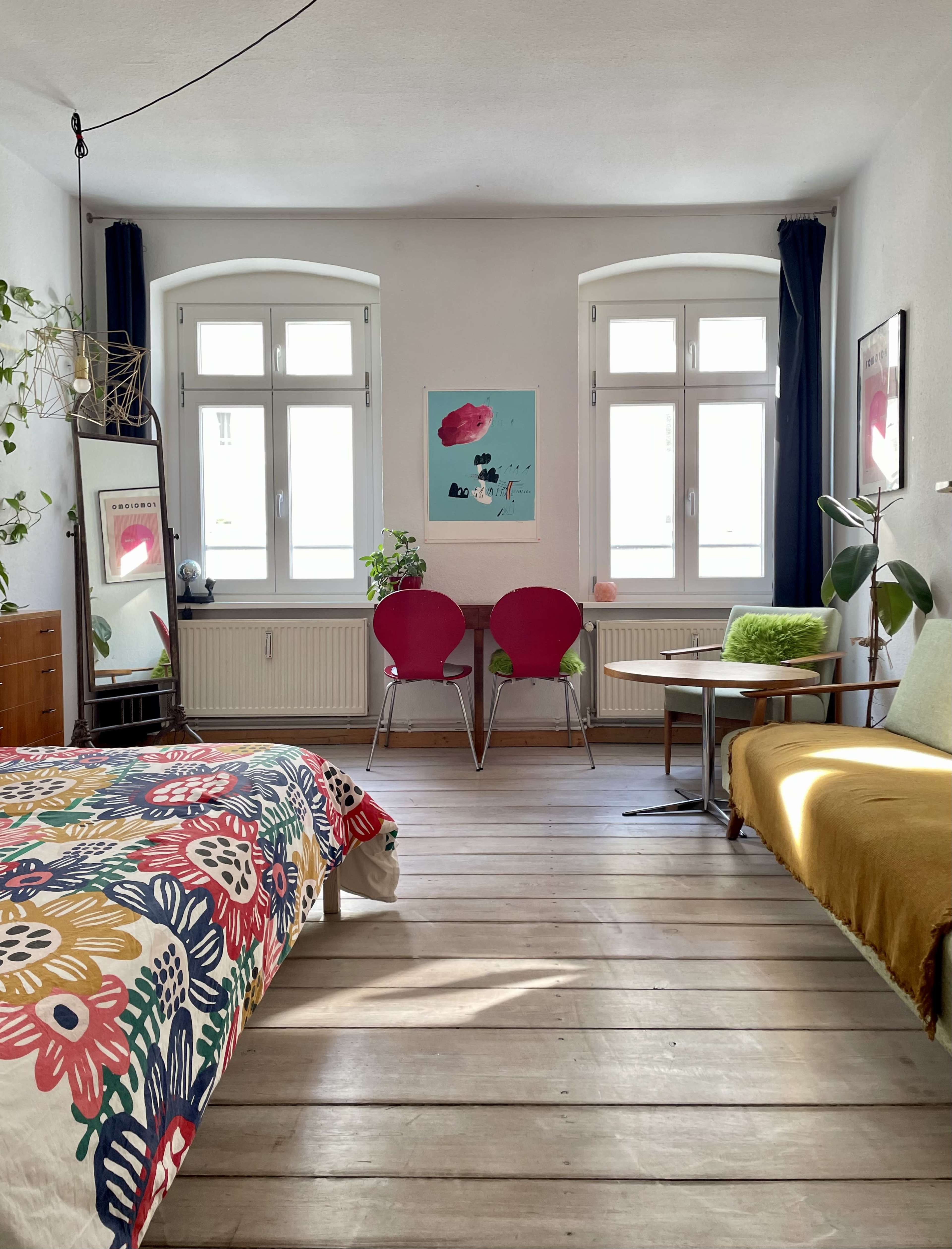 A brightly lit room features a bed with a colorful floral cover, a small table with two pink chairs, and a couch against the wall, surrounded by large windows and indoor plants.