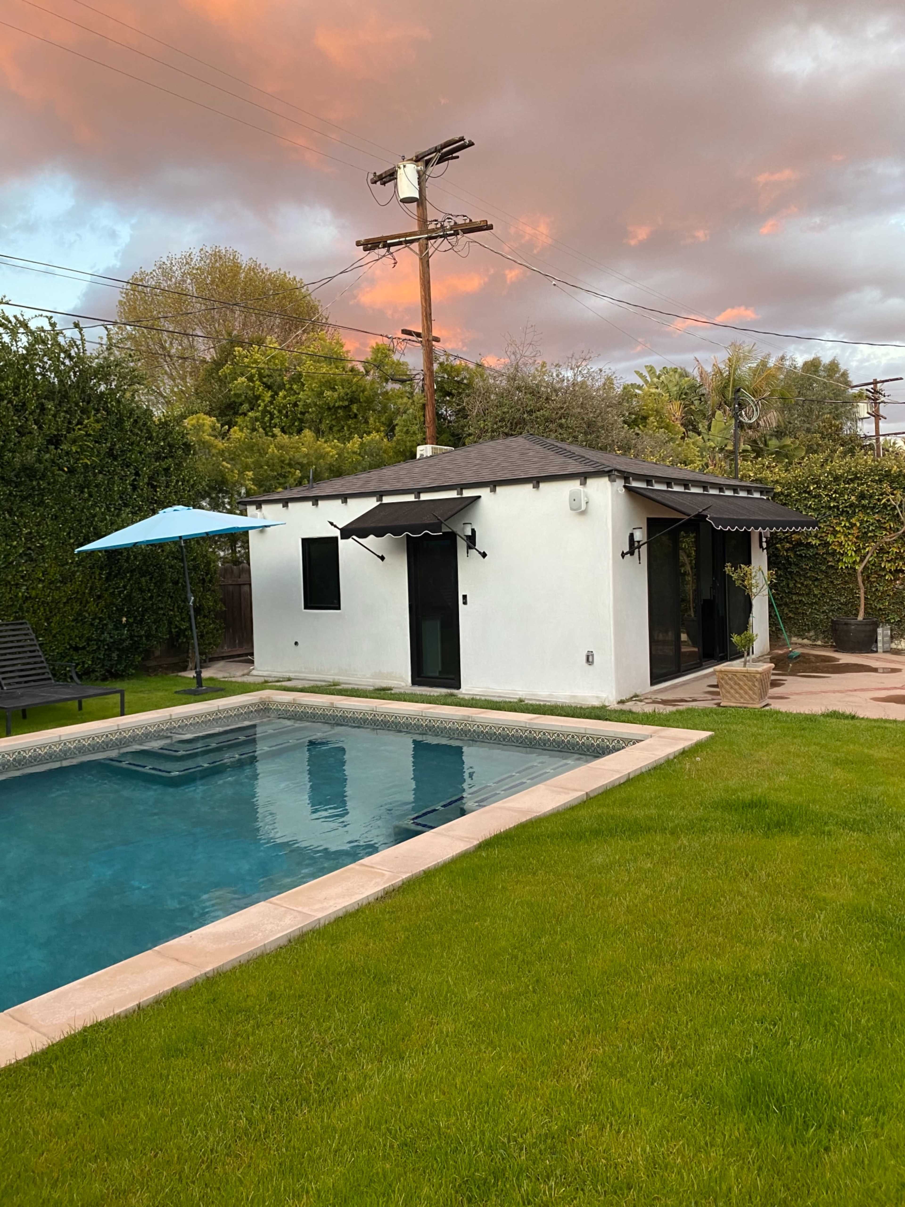 A small white structure with a dark roof sits beside a swimming pool in a well-kept lawn, with an umbrella providing shade nearby and trees in the background under a cloudy sky.