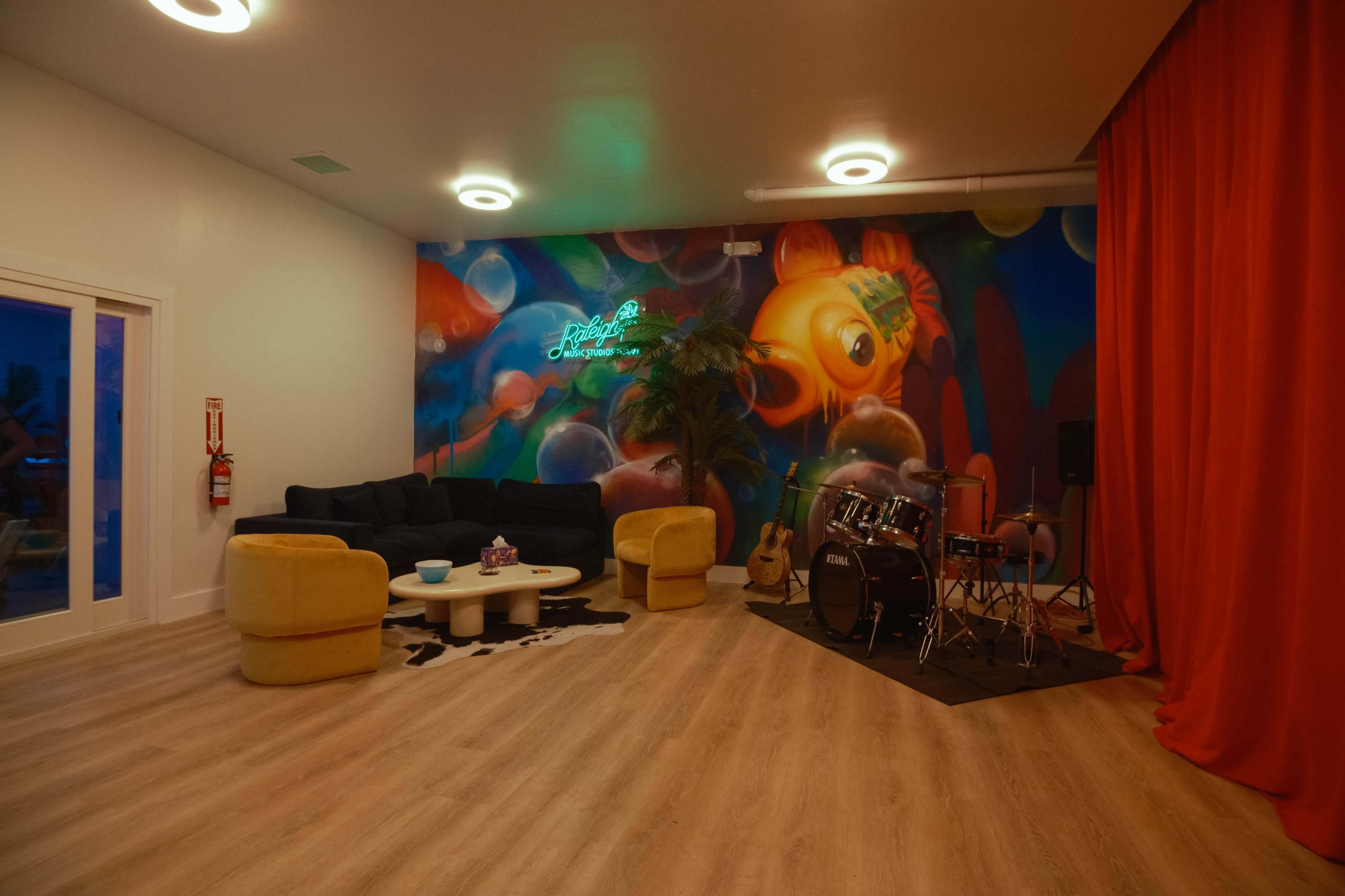 The interior features a lounge area with a dark couch, yellow chairs, a coffee table, and a mural on the wall alongside a small stage with a drum set.