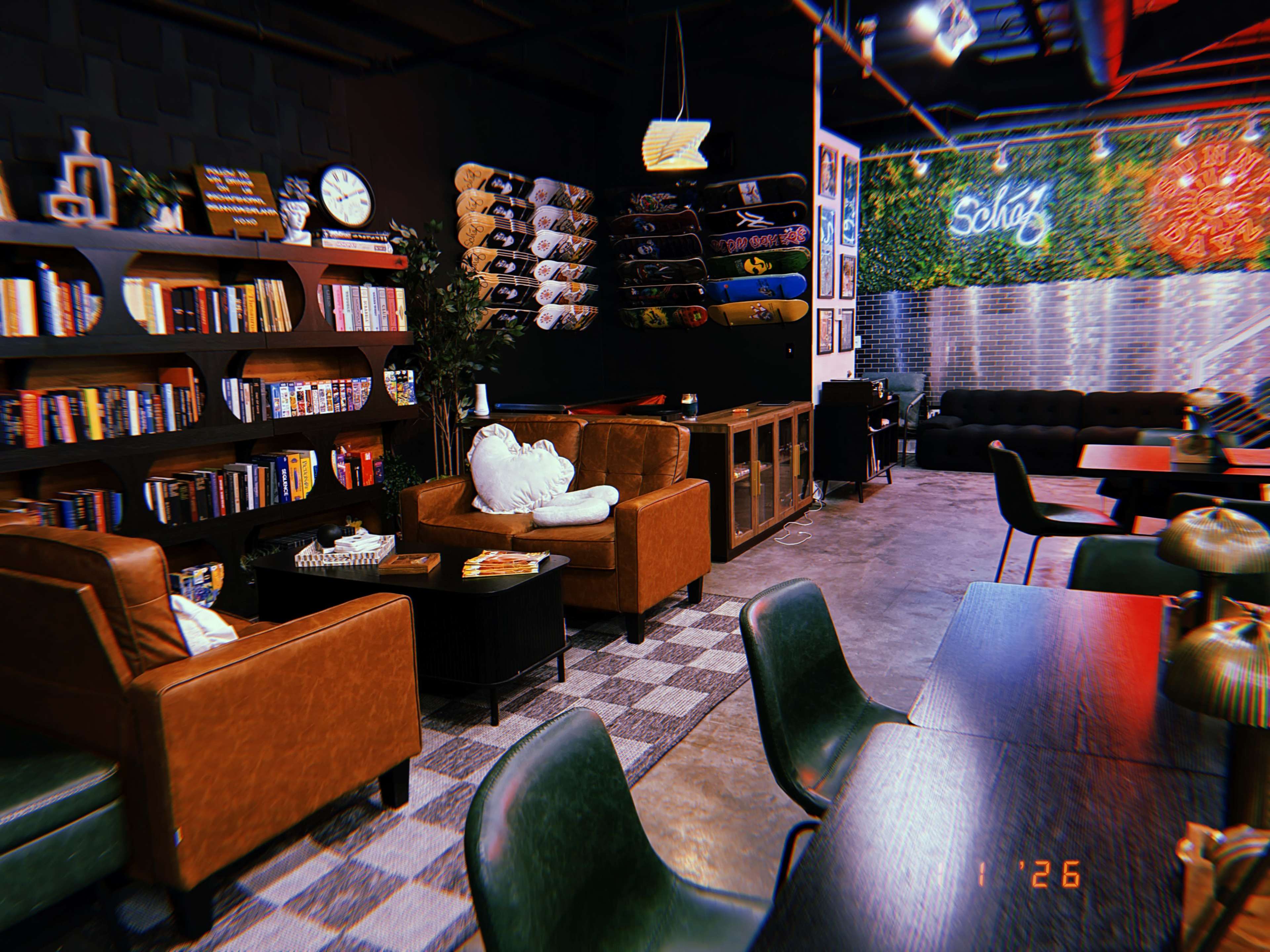 The image shows a modern café interior featuring brown leather furniture, shelves lined with books, skateboards displayed on the wall, and lush greenery.