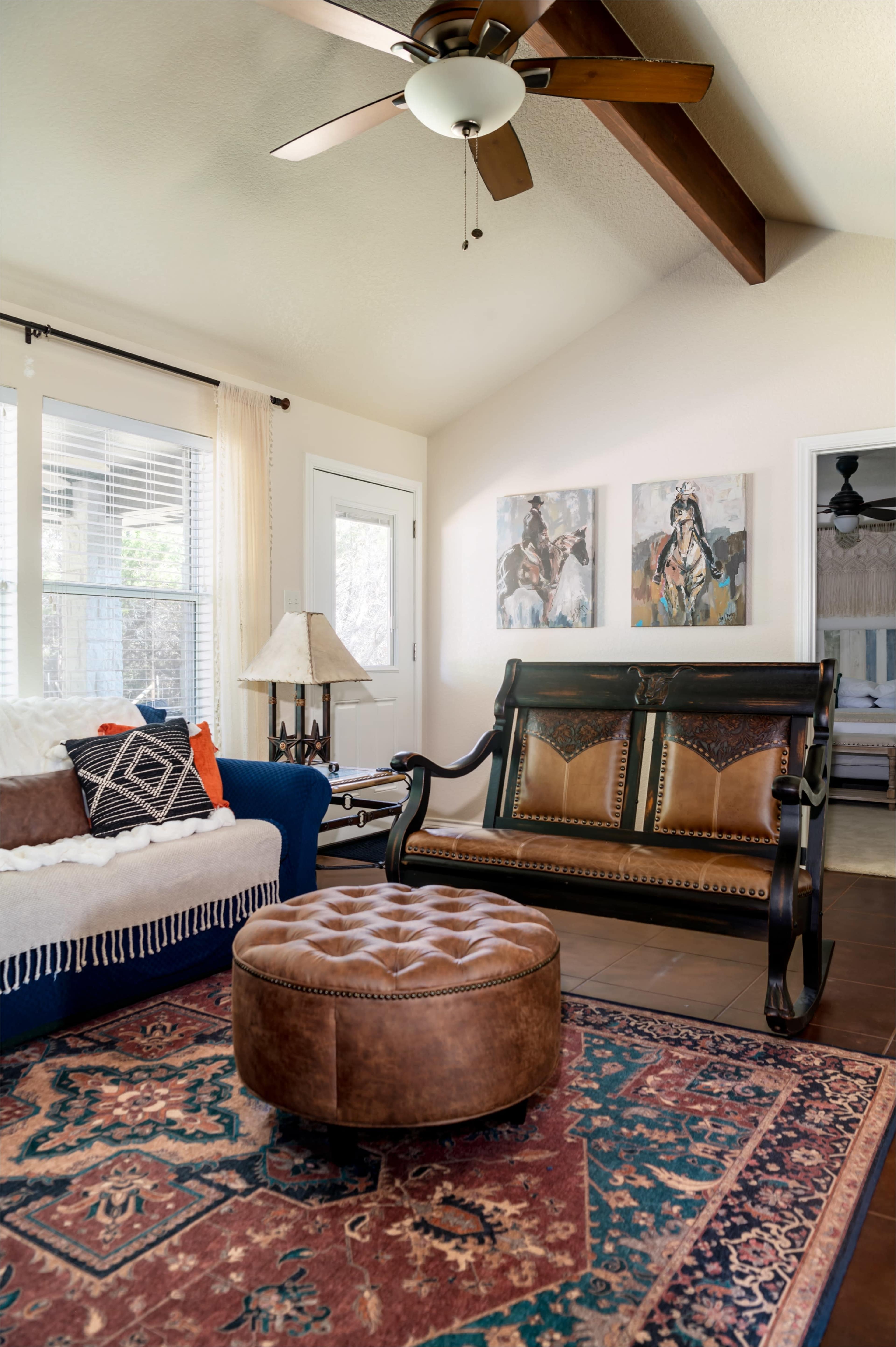 A living room features a blue couch, a wooden rocking chair, a round ottoman, and artwork on the walls.