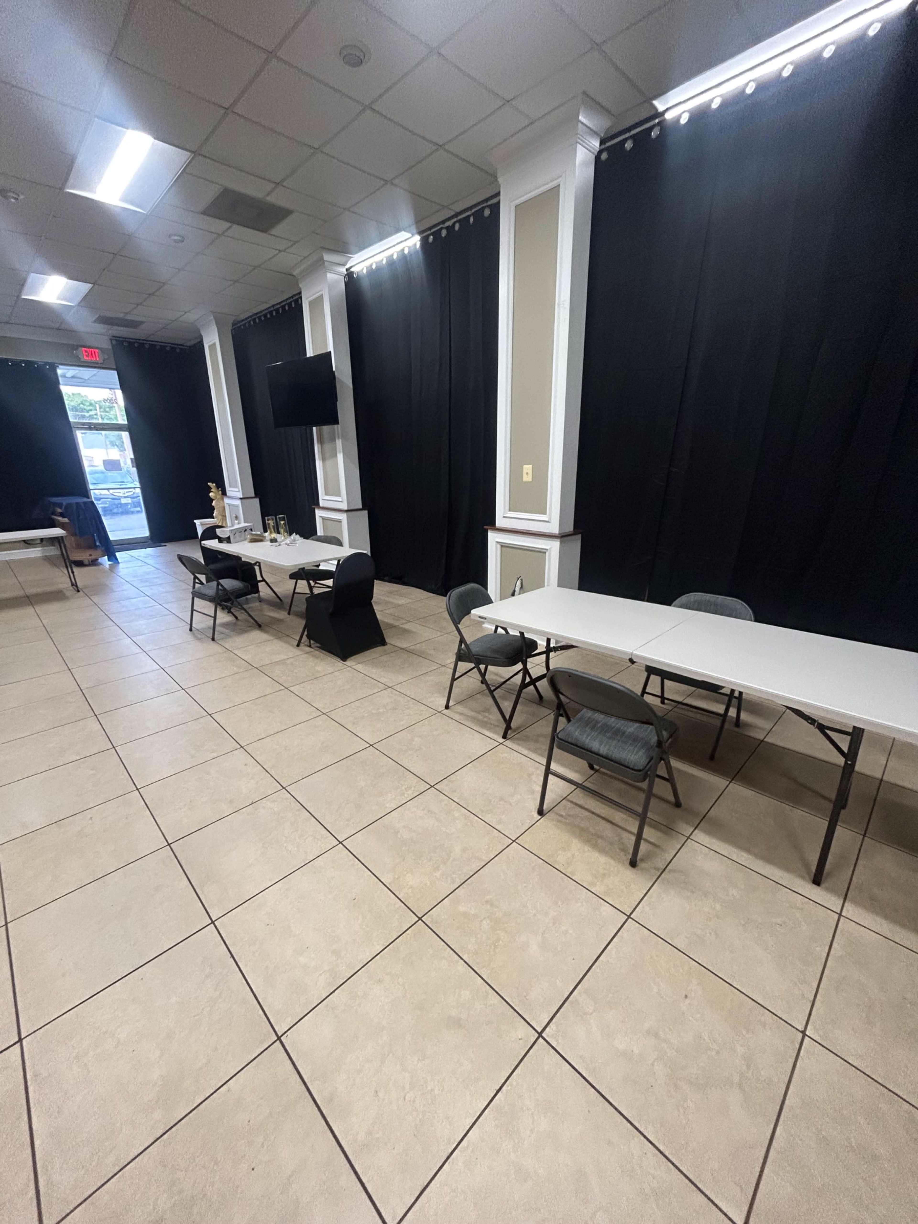An empty room with white folding tables and chairs arranged on tiled flooring, flanked by black curtains and a wall-mounted television.