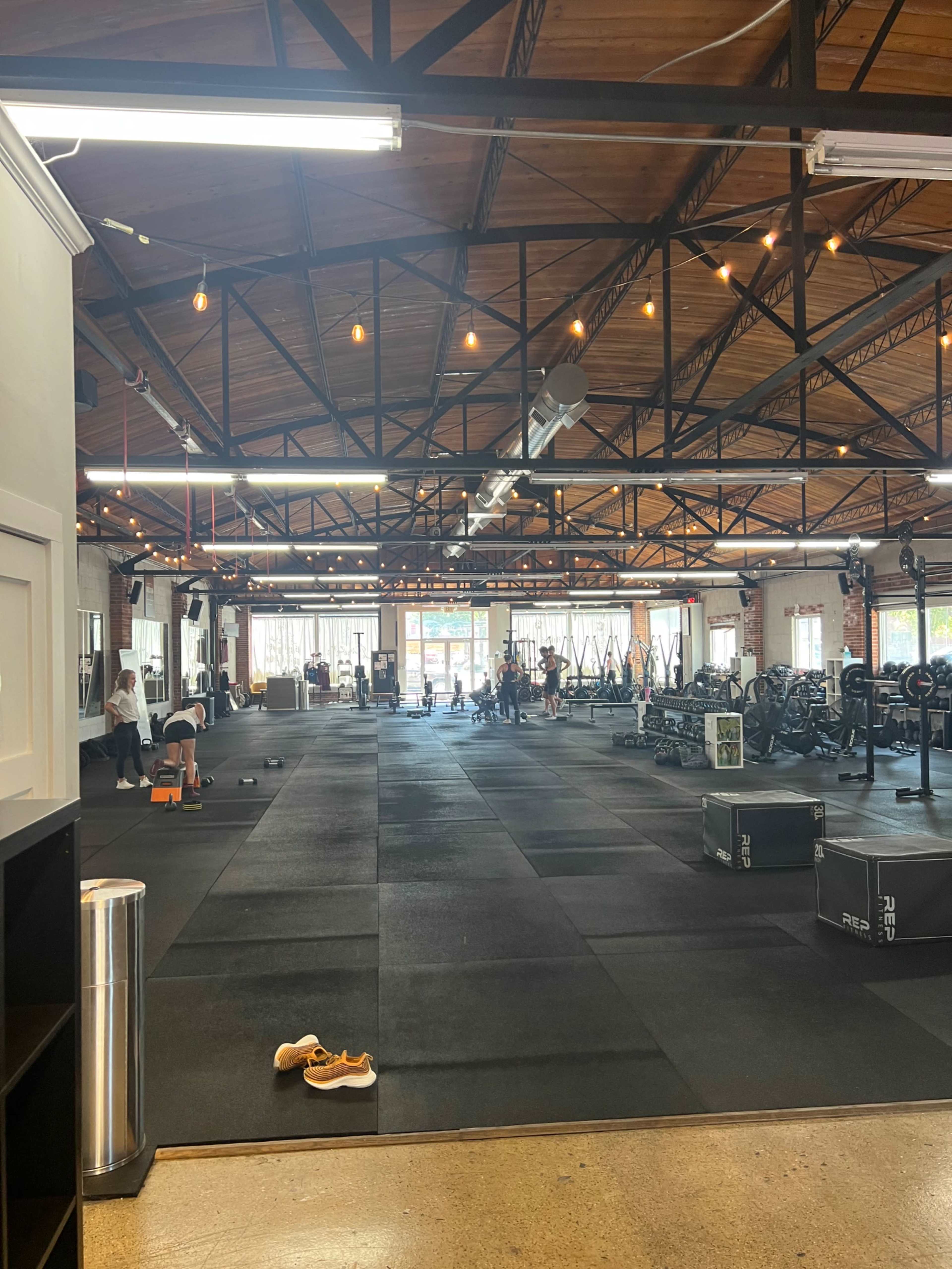 The image shows a spacious gym interior with black rubber flooring, weightlifting equipment, and several people engaged in workouts.