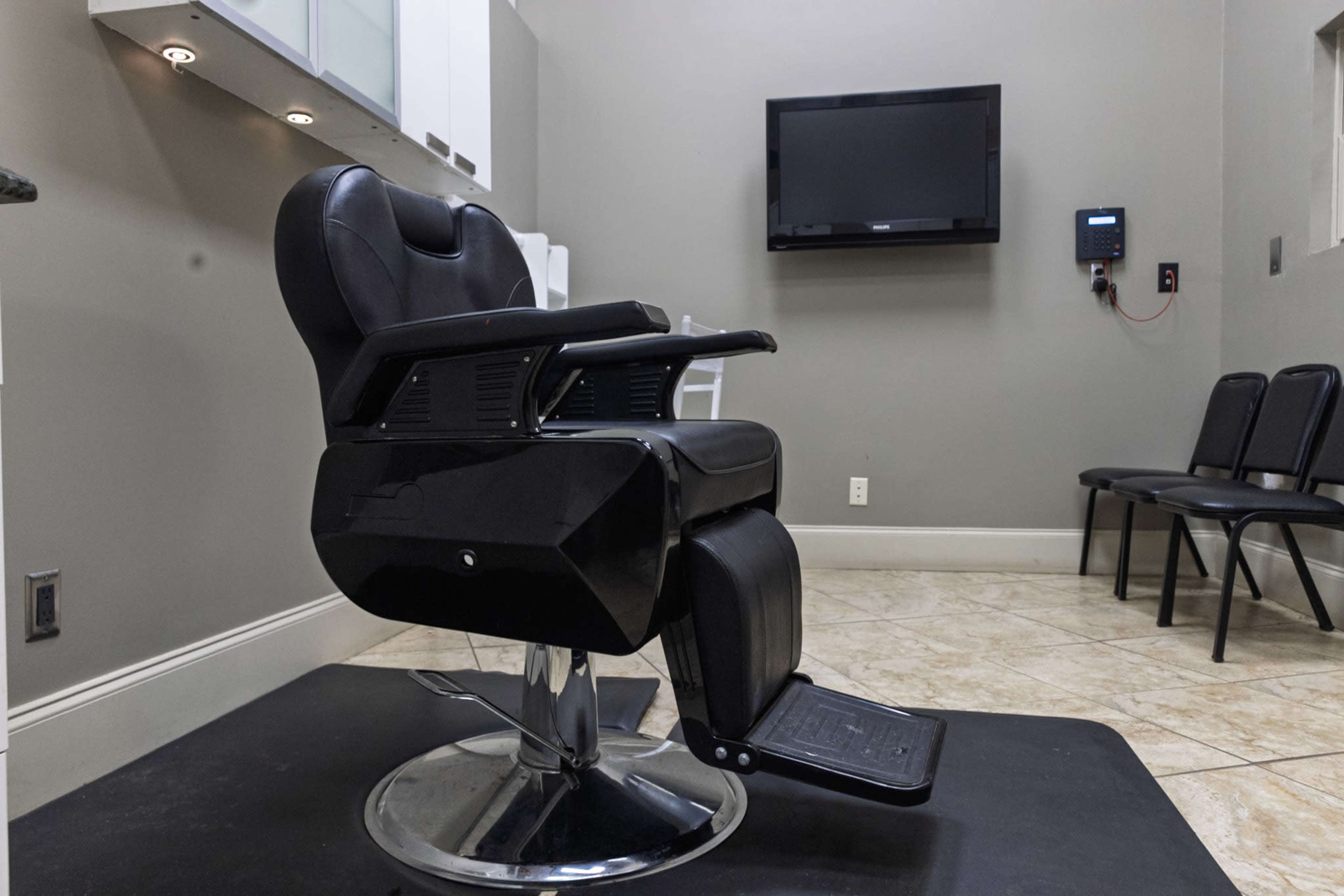 The image shows a black barber chair positioned in a hair salon with a wall-mounted television and empty chairs in the background.