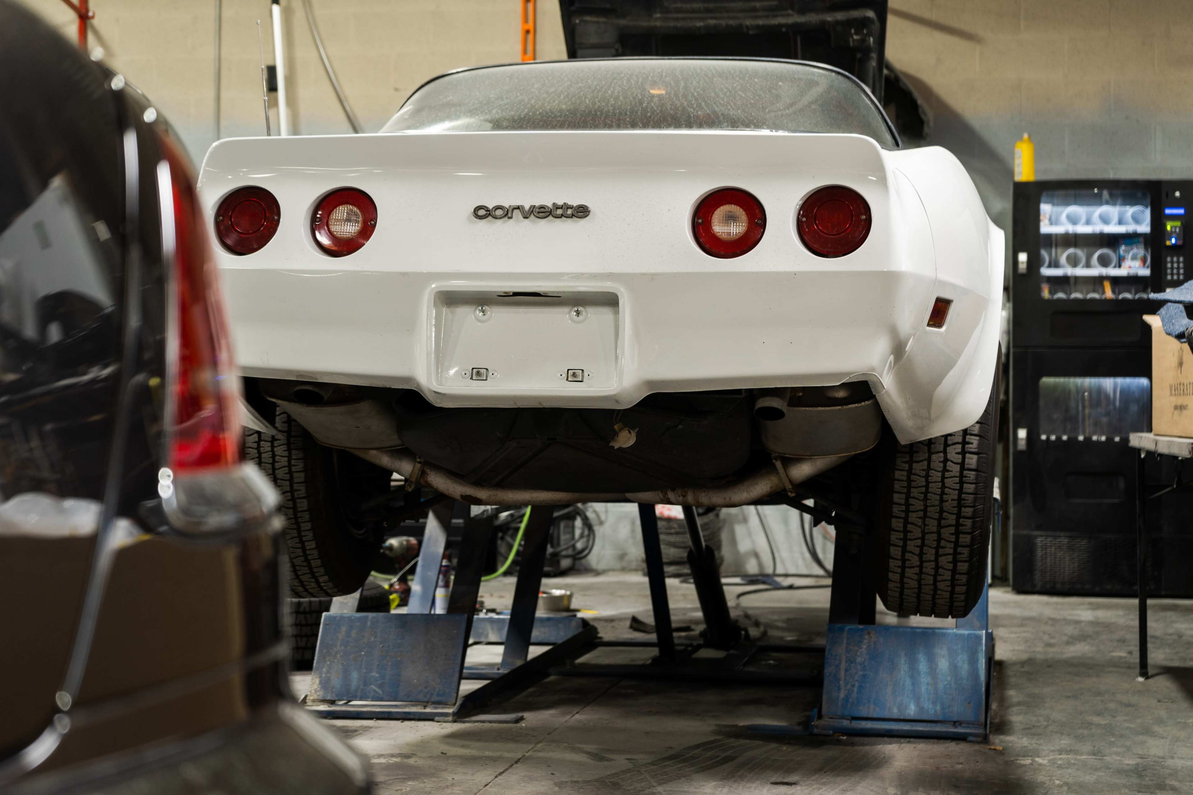 A white Corvette is elevated on a pair of blue ramps in a garage.