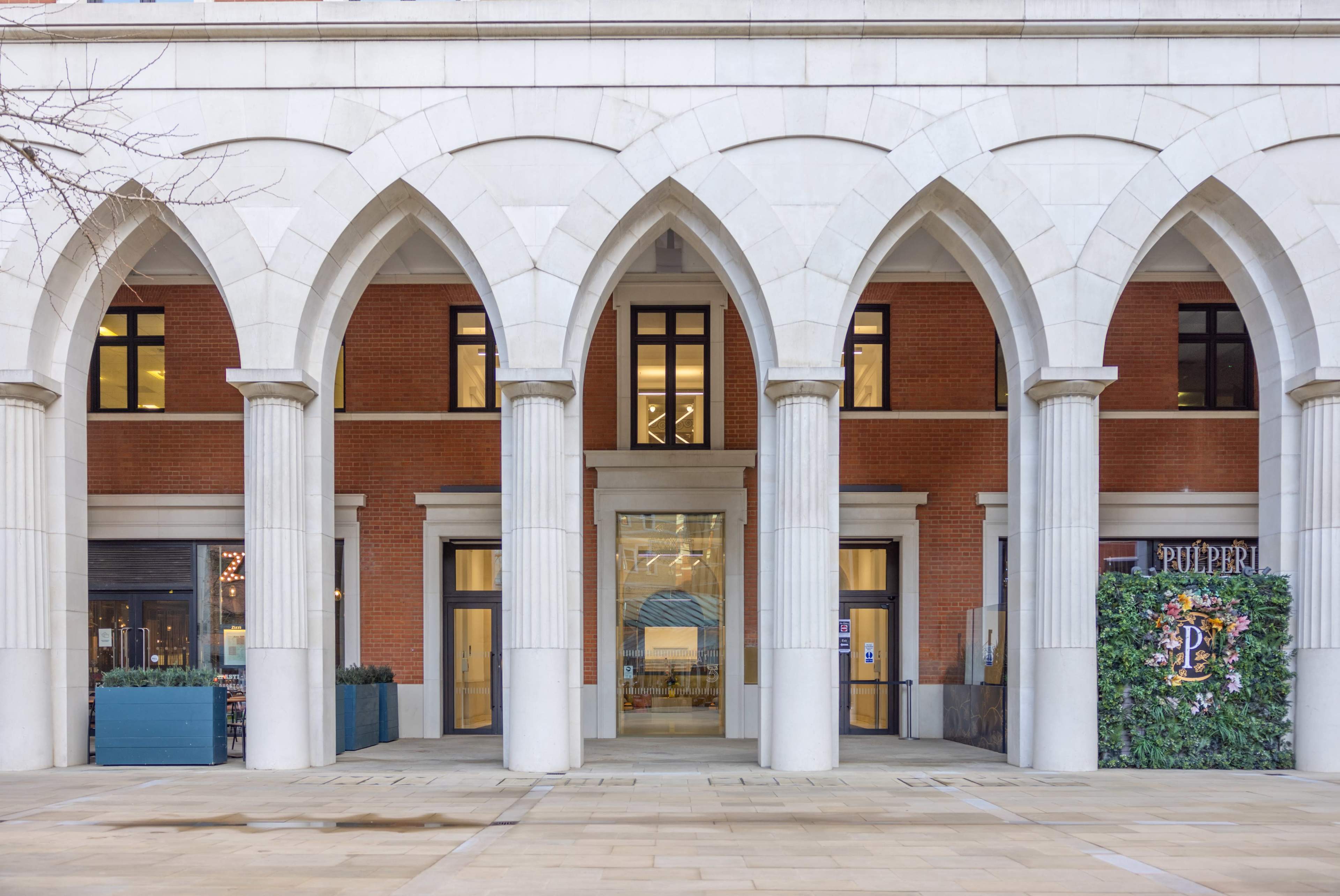 An architectural facade with multiple arches, solid columns, and a combination of brick and stone elements, leading to a glass entrance.