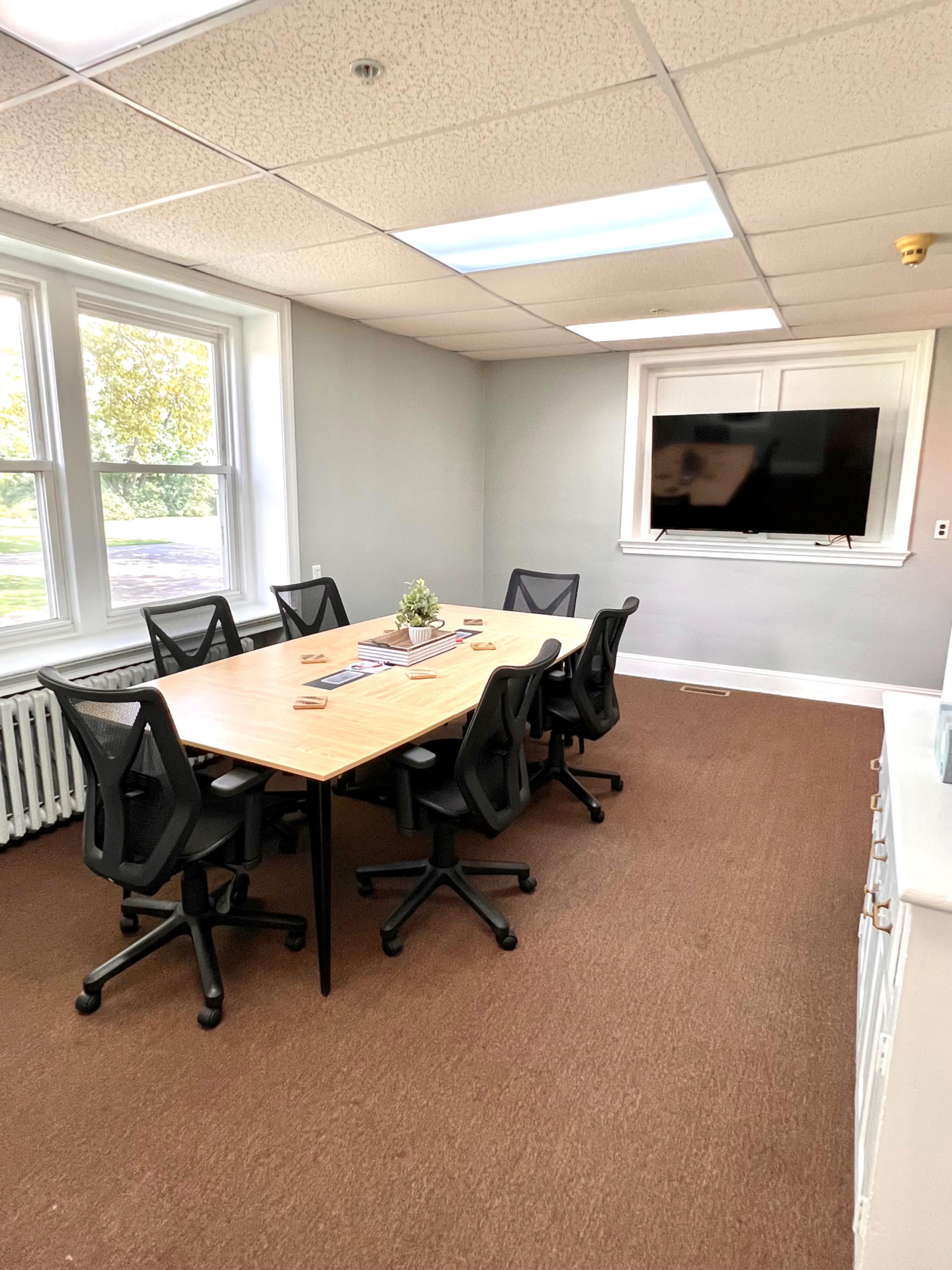 A meeting room with a rectangular table surrounded by six black office chairs and a mounted television on the wall.