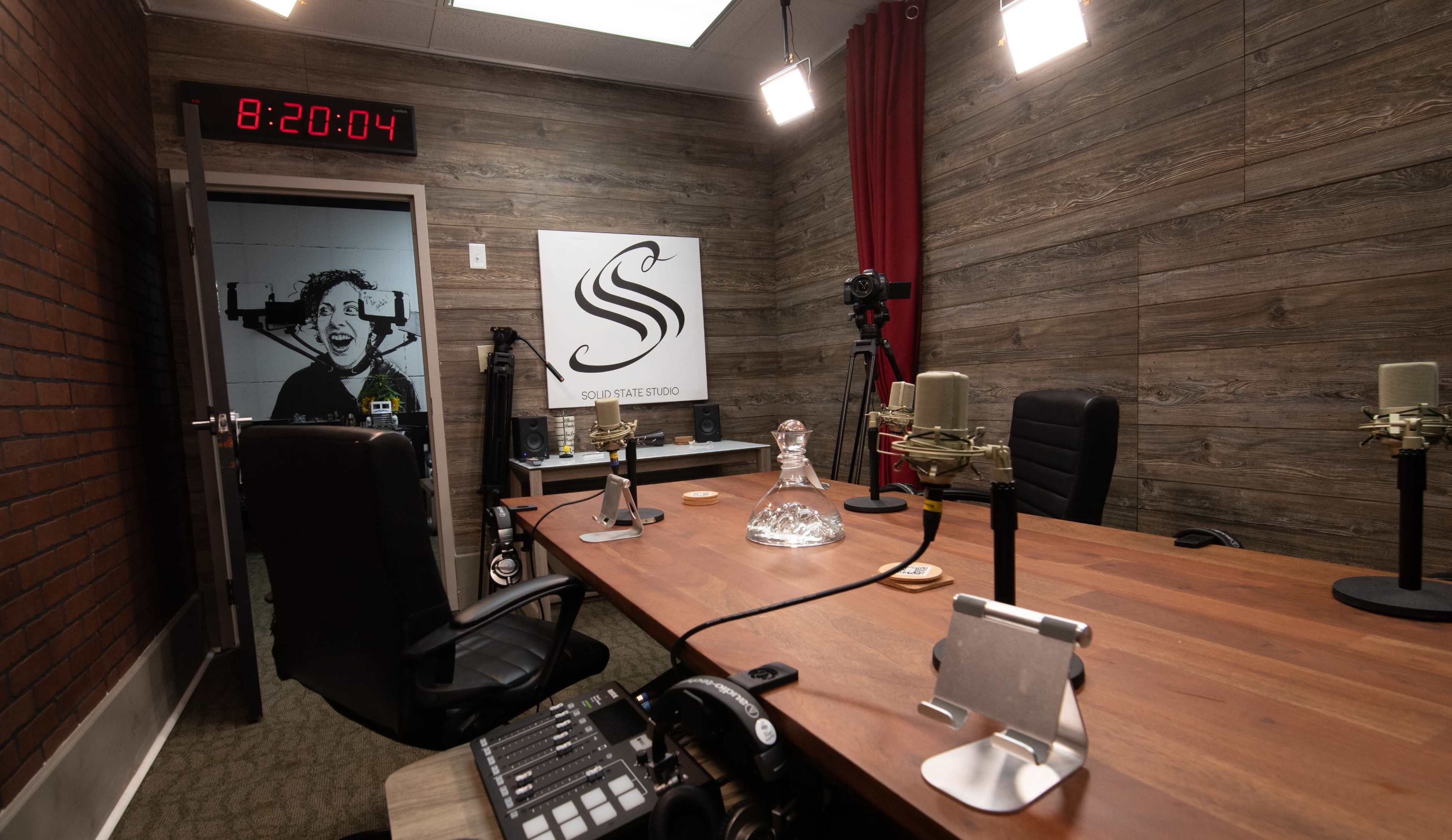 The image shows a recording studio with a wooden table, microphones, and a large clock displaying the time.
