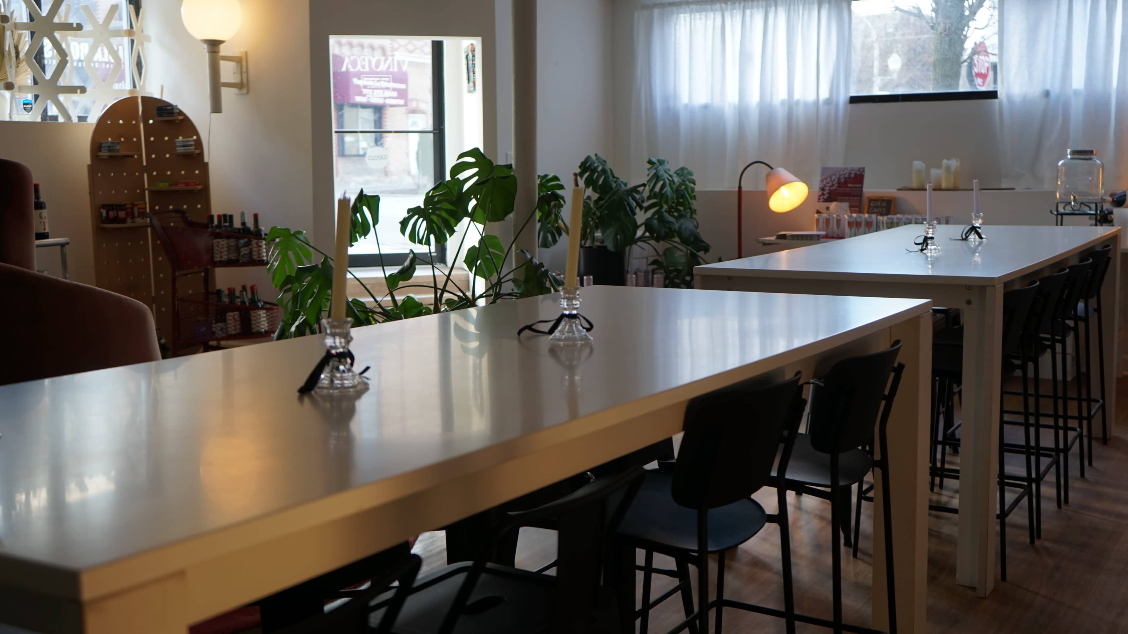 The image shows a spacious interior with long white tables, black chairs, and several green plants, illuminated by natural light coming through large windows.