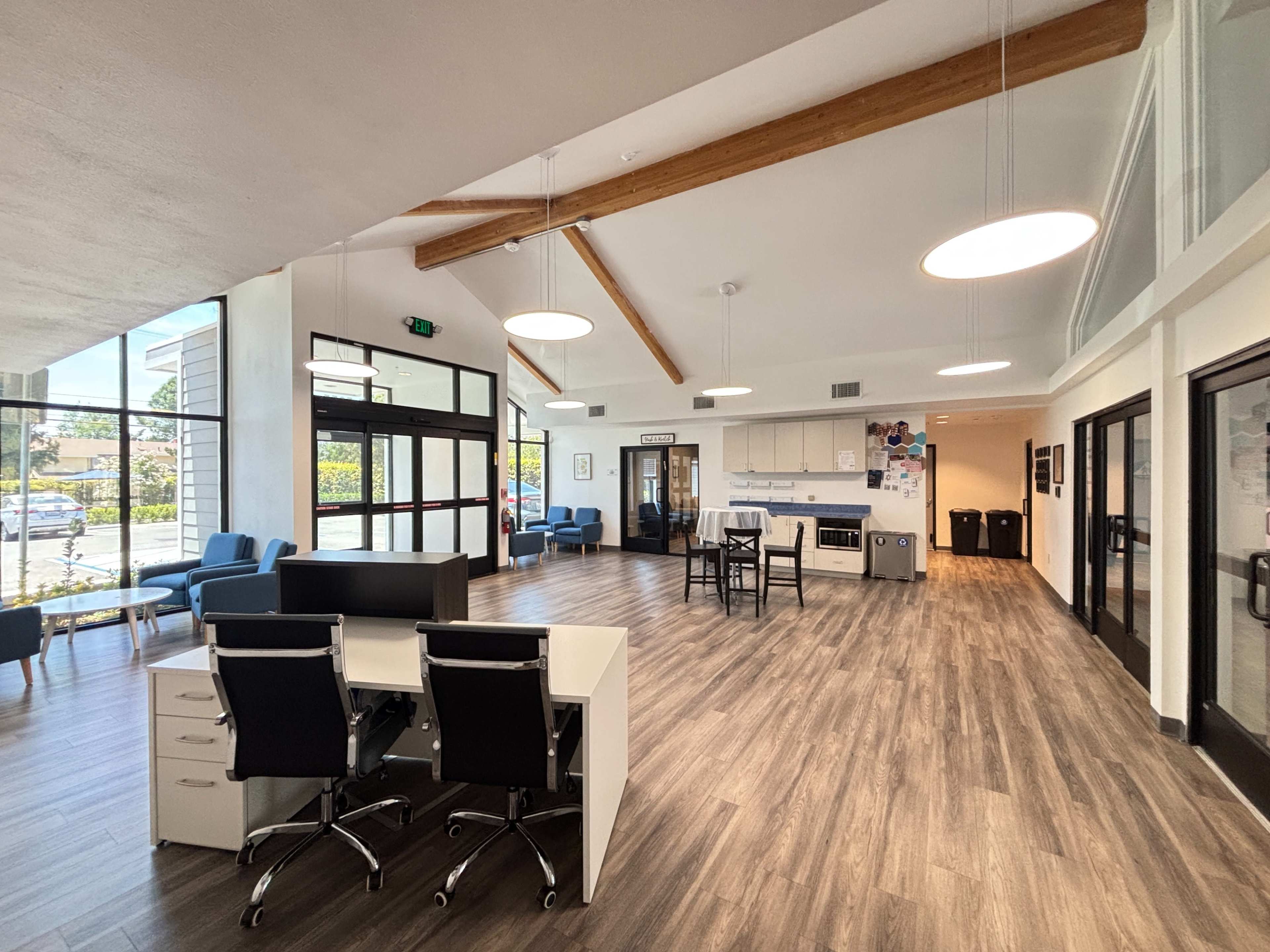The image shows a spacious interior of a modern office or community space with wooden beams, a kitchenette, and seating areas.