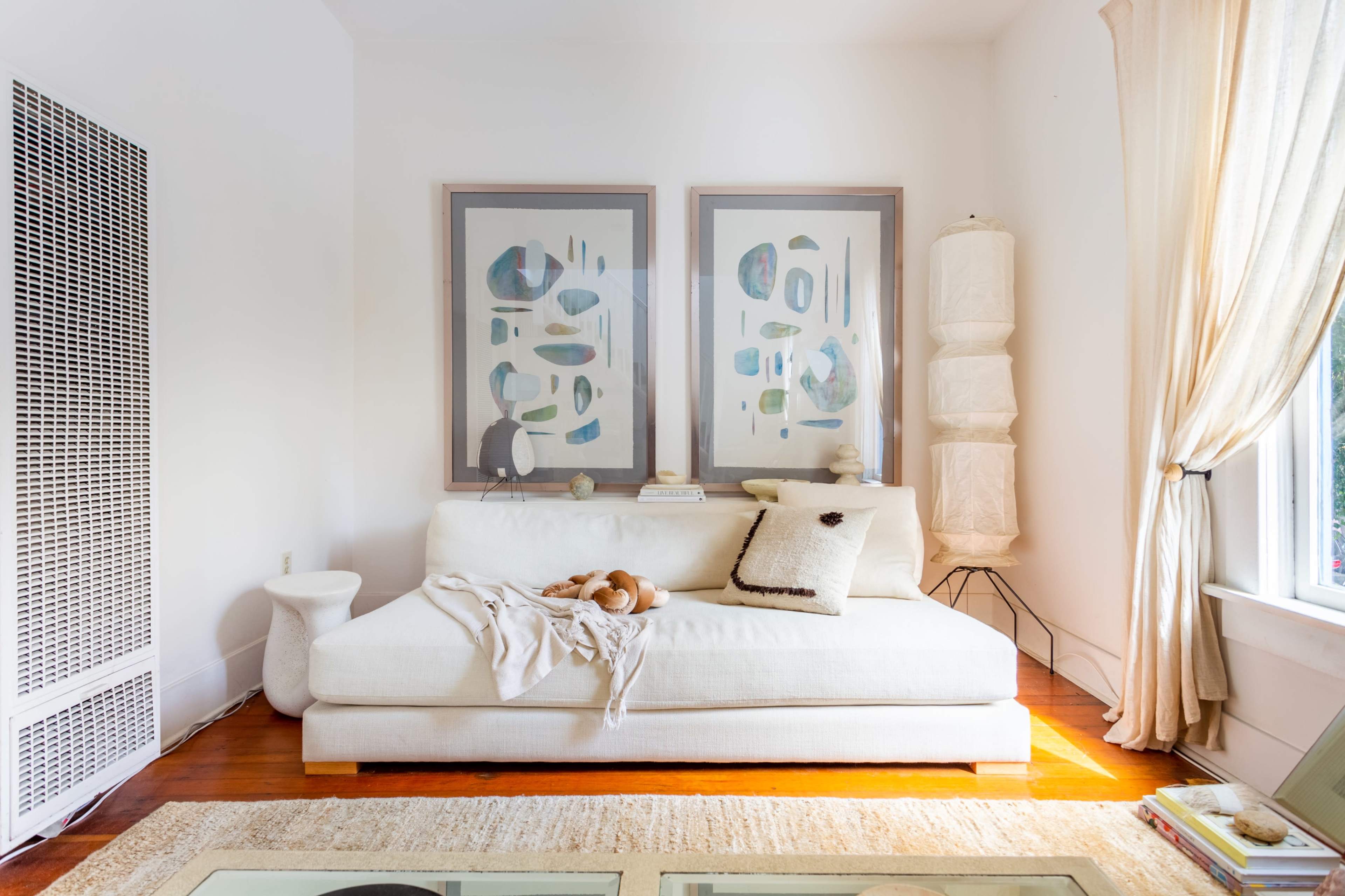 A bright, airy living room with a white sofa, two framed abstract paintings on the wall, and a light-colored floor lamp beside a window.
