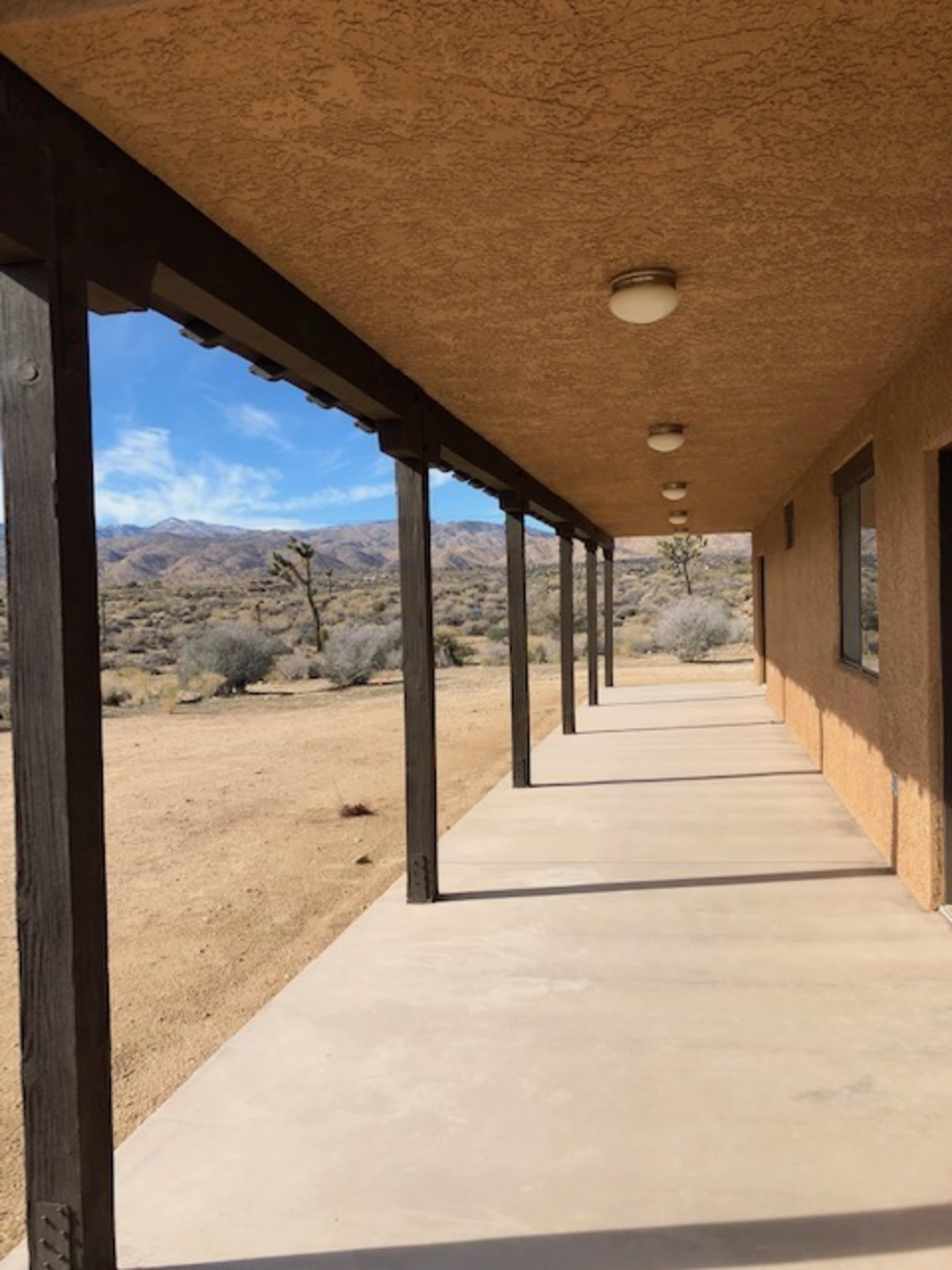 A long, shadowy walkway with a flat concrete surface and a series of lights along the ceiling, leading towards a desert landscape with sparse vegetation and mountains in the background.